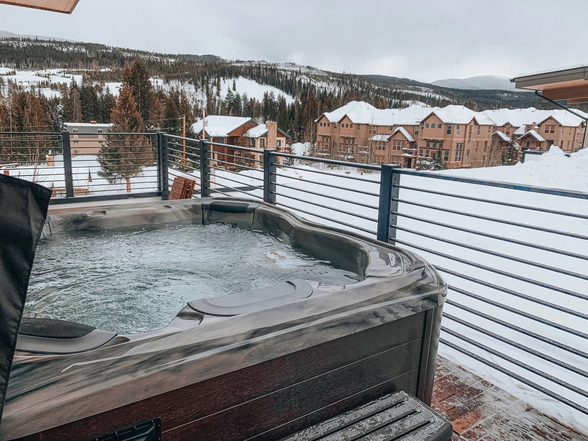 Outdoor hot tub on a balcony overlooking snow-covered mountain village and forested hills.