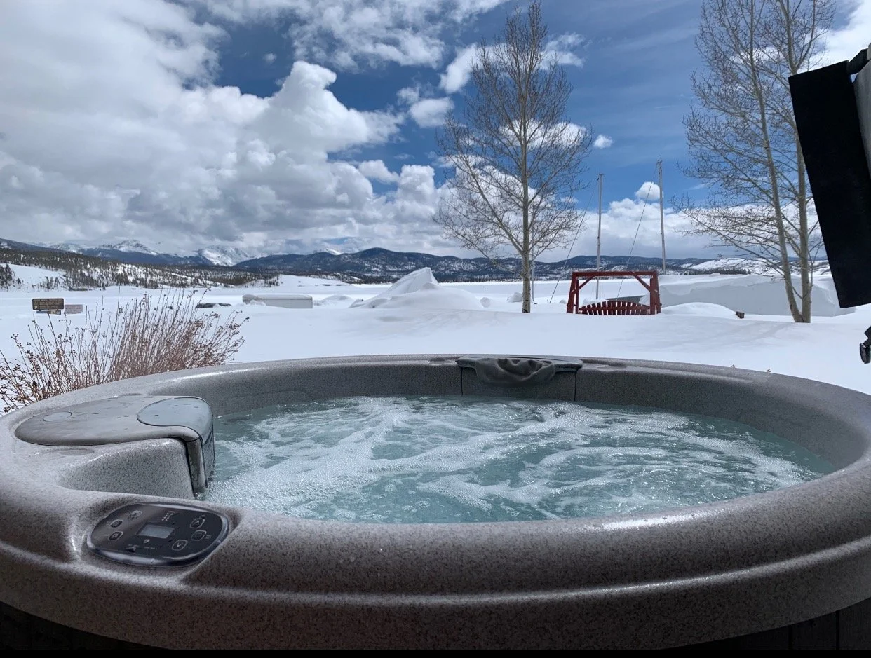 A hot tub outdoors in a snowy landscape with snow-covered mountains, leafless trees, and a blue sky with clouds.