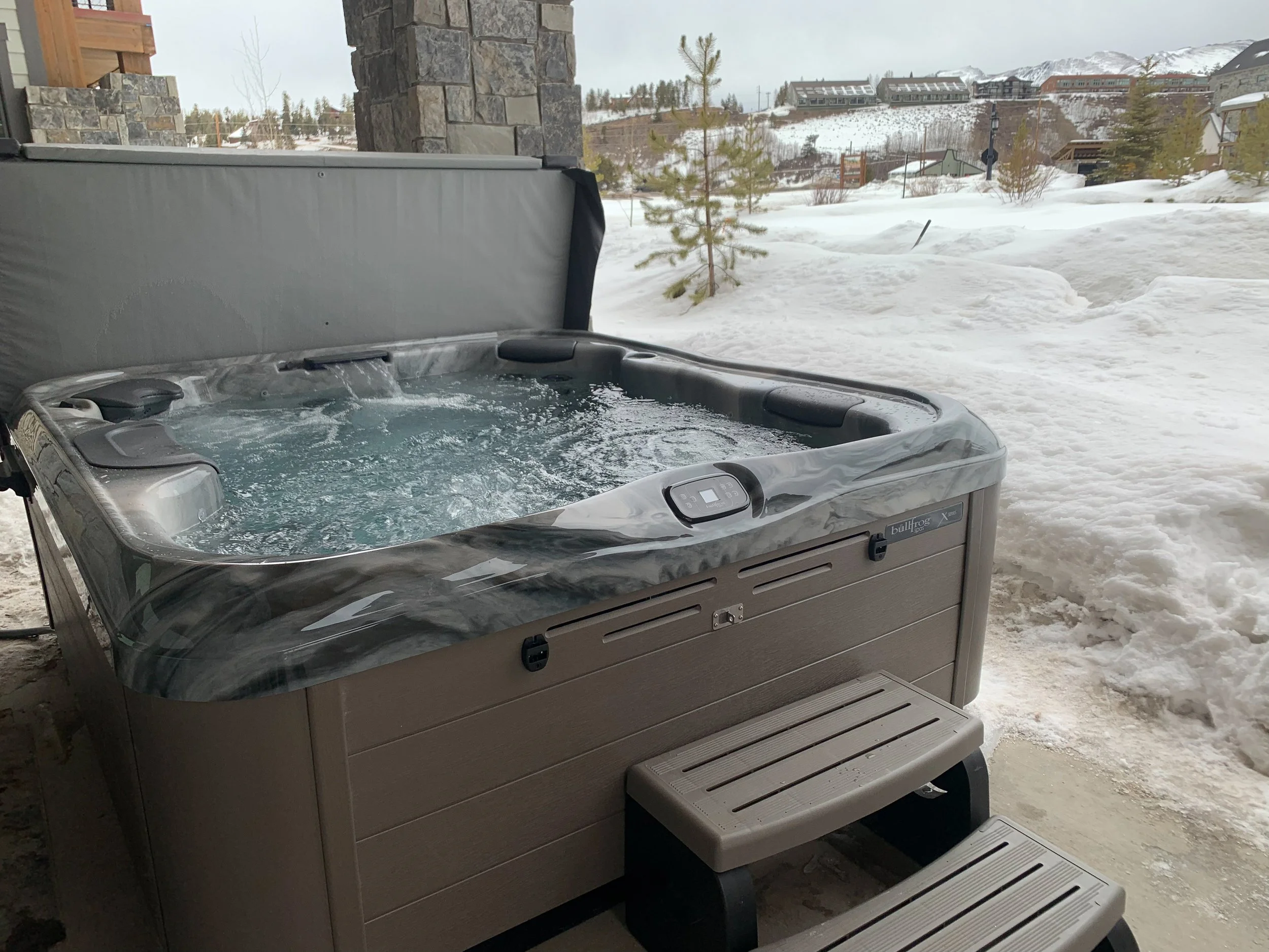 An outdoor hot tub with water and bubbles on a snowy porch, with snow-covered hills and houses in the background.
