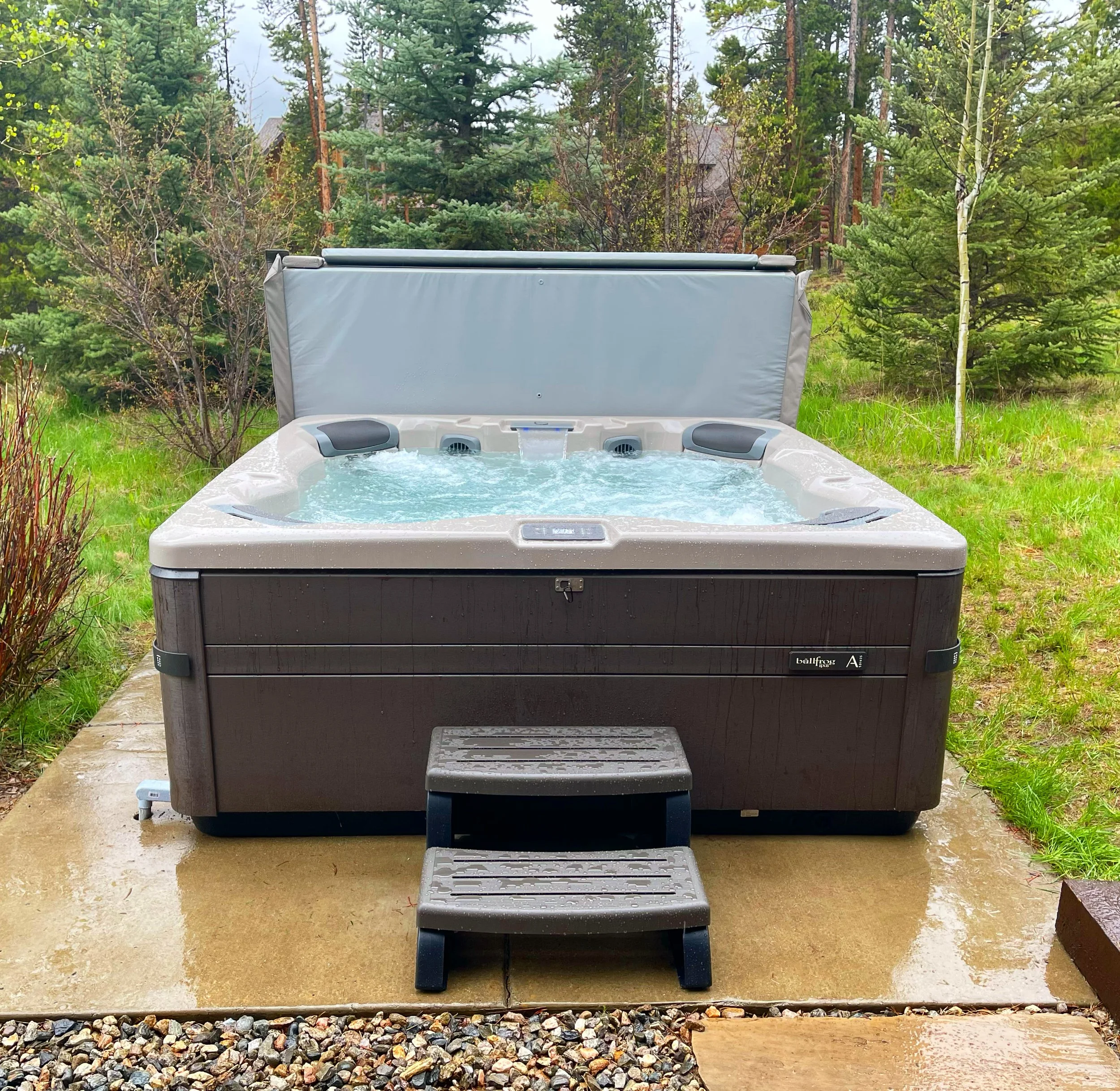 Outdoor hot tub with water jets on a concrete patio, surrounded by green grass and trees in a forested setting.