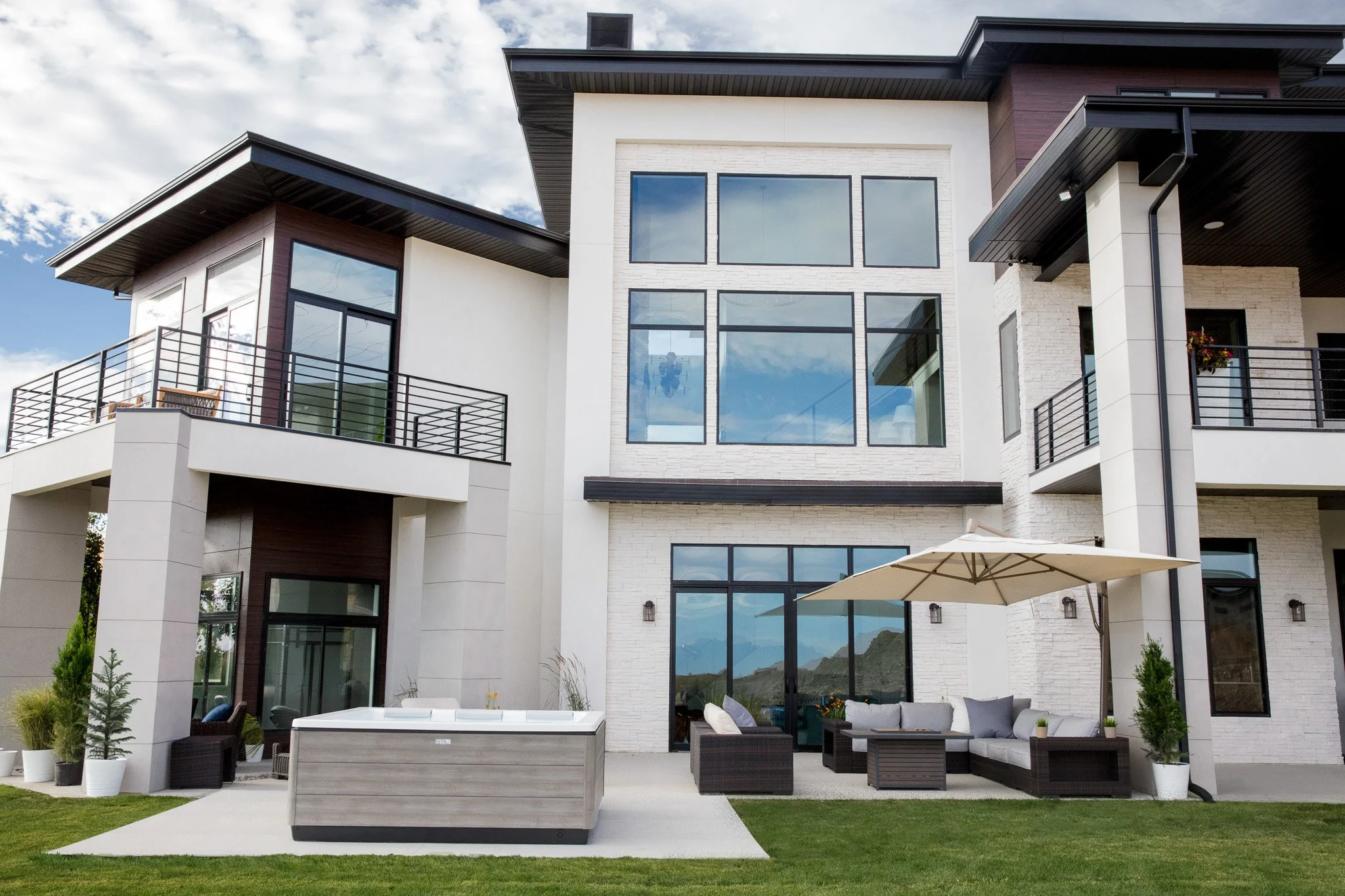 Modern multi-story house with large windows, outdoor patio furniture, an umbrella, potted plants, and a hot tub on a grassy lawn under a partly cloudy sky.