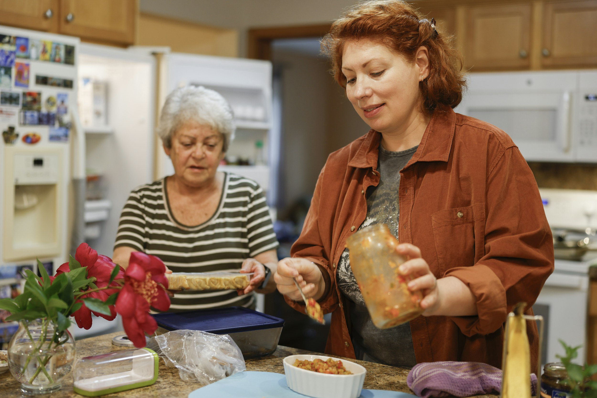 Two women in a kitchen preparing food. One woman with short gray hair wears a striped shirt, and the other woman with curly red hair wears an orange jacket. The table has flowers, bowls of food, and kitchen items.