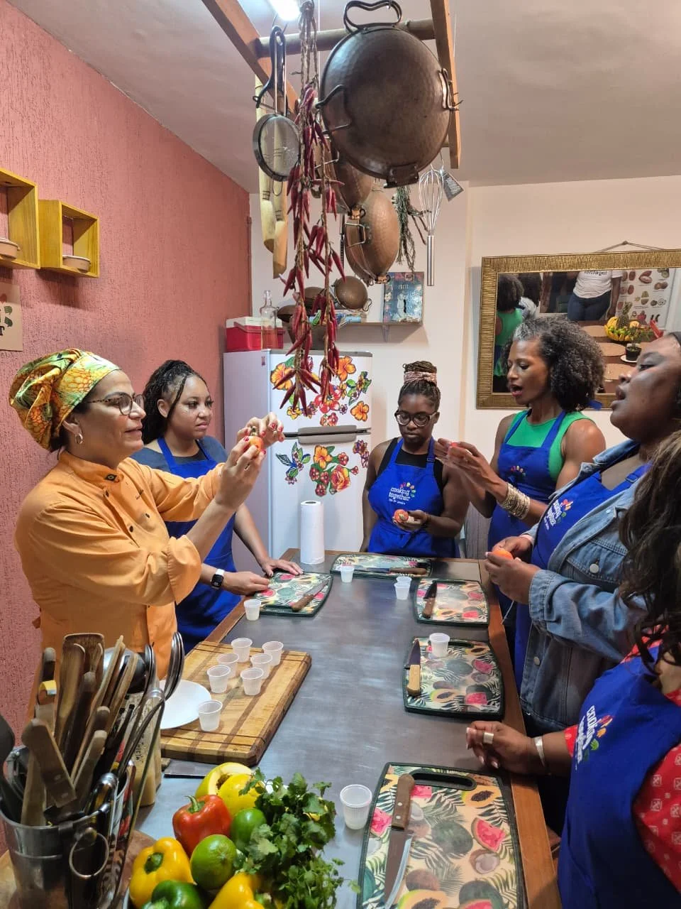 A group of women in a kitchen participating in a cooking class or demonstration. The woman on the left is wearing an orange shirt and a colorful headwrap, holding a small orange item. The other women are wearing blue aprons, and one woman in the cent