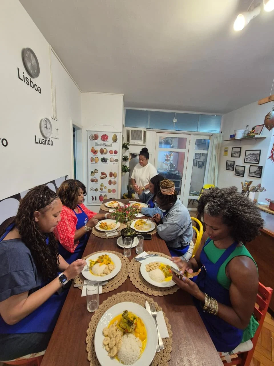 Six women sitting around a dining table with plates of rice, vegetables, and meat, while one woman stands and prepares food in the background inside a cozy dining room.
