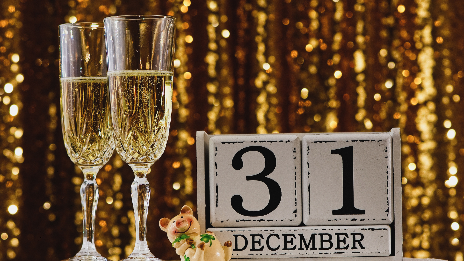 Two glasses of champagne, a small pig figurine, and a calendar showing December 31st, with a shimmering golden background.