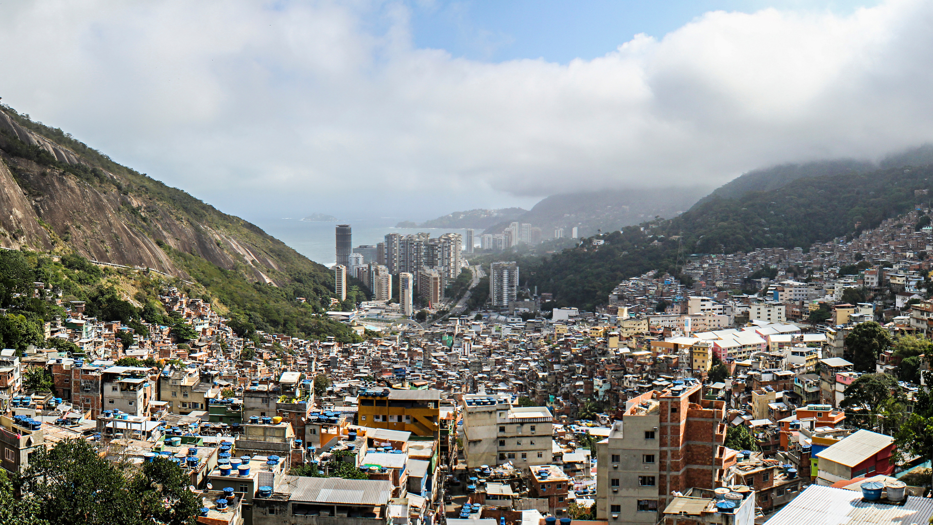 A cityscape with densely packed buildings on a hillside, tall modern apartment buildings in the middle, mountains on either side, and cloudy sky overhead.