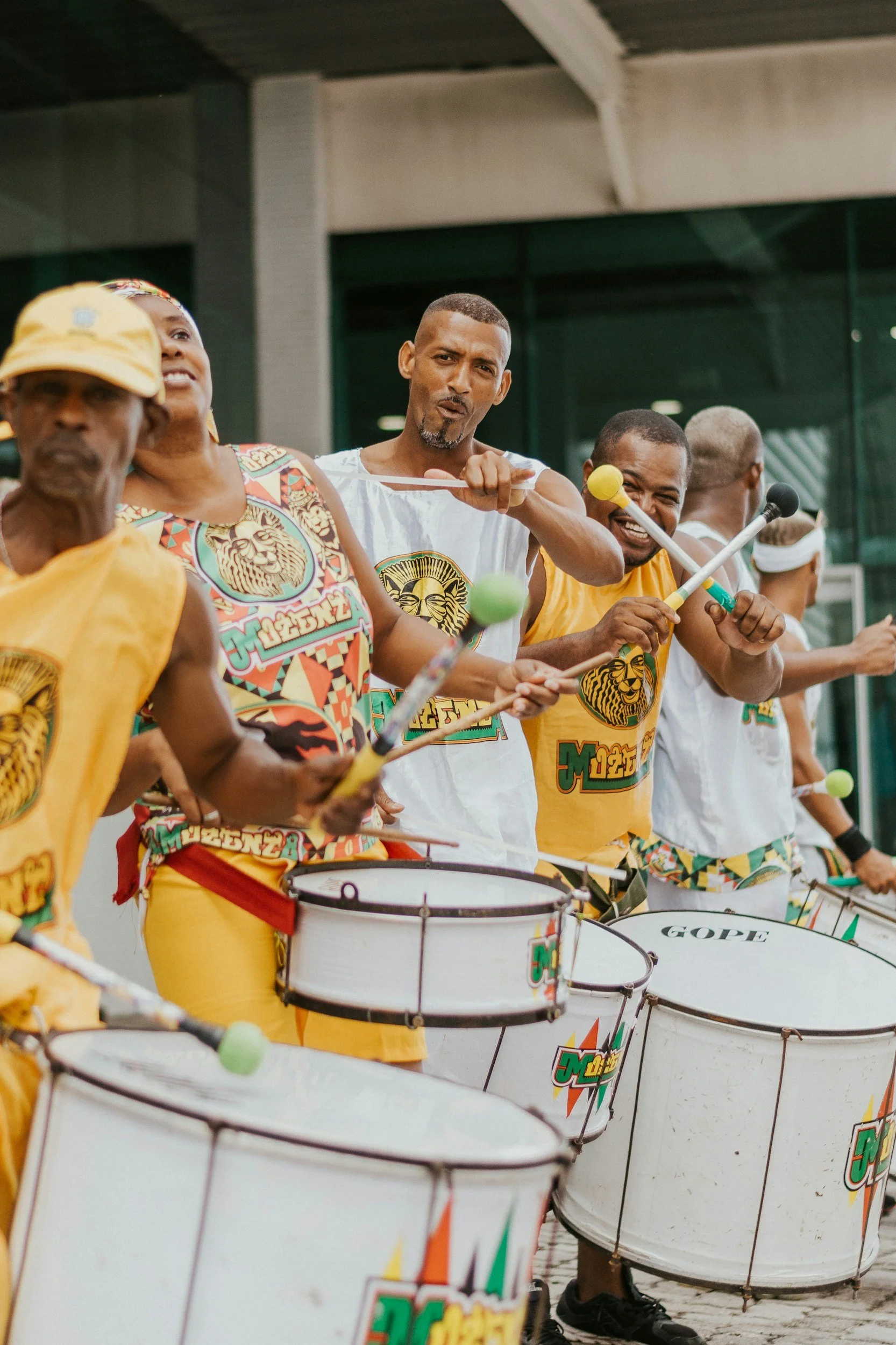 A group of musicians playing drums and percussion instruments outdoors, wearing colorful shirts with a lion logo, some smiling and some focused.