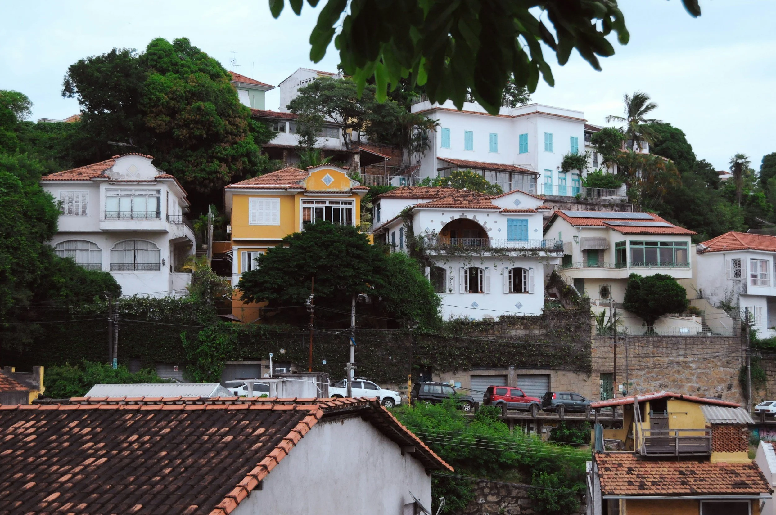 Houses stacked on a hillside with trees, parked cars, and a tiled roof in foreground, with cloudy sky above.