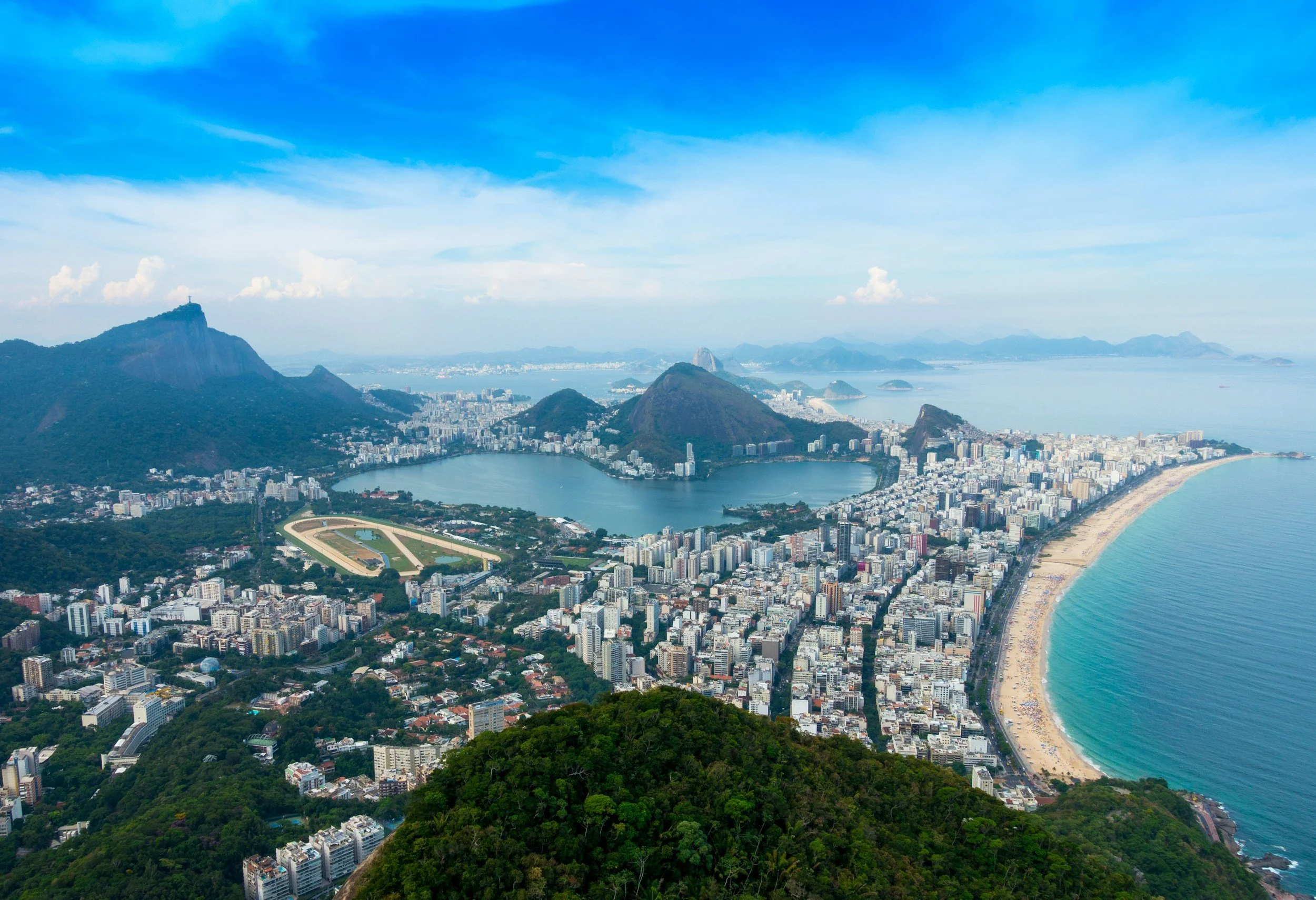 A panoramic aerial view of Rio de Janeiro, Brazil, showing the cityscape, beaches, and iconic mountains including Sugarloaf Mountain and Corcovado, under a blue sky with clouds.