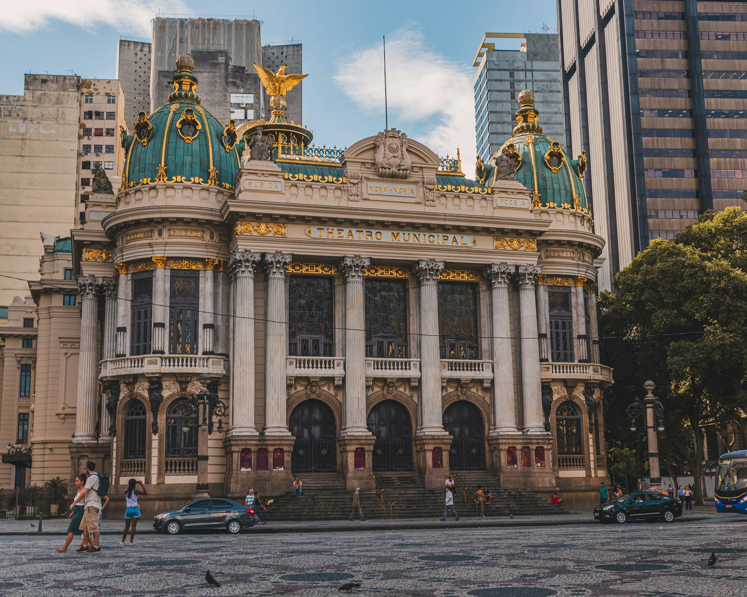 A historic theater building with ornate architecture, columns, and green domes with gold accents, located in a city with modern skyscrapers in the background. Pedestrians and cars are visible in the street in front of the theater.