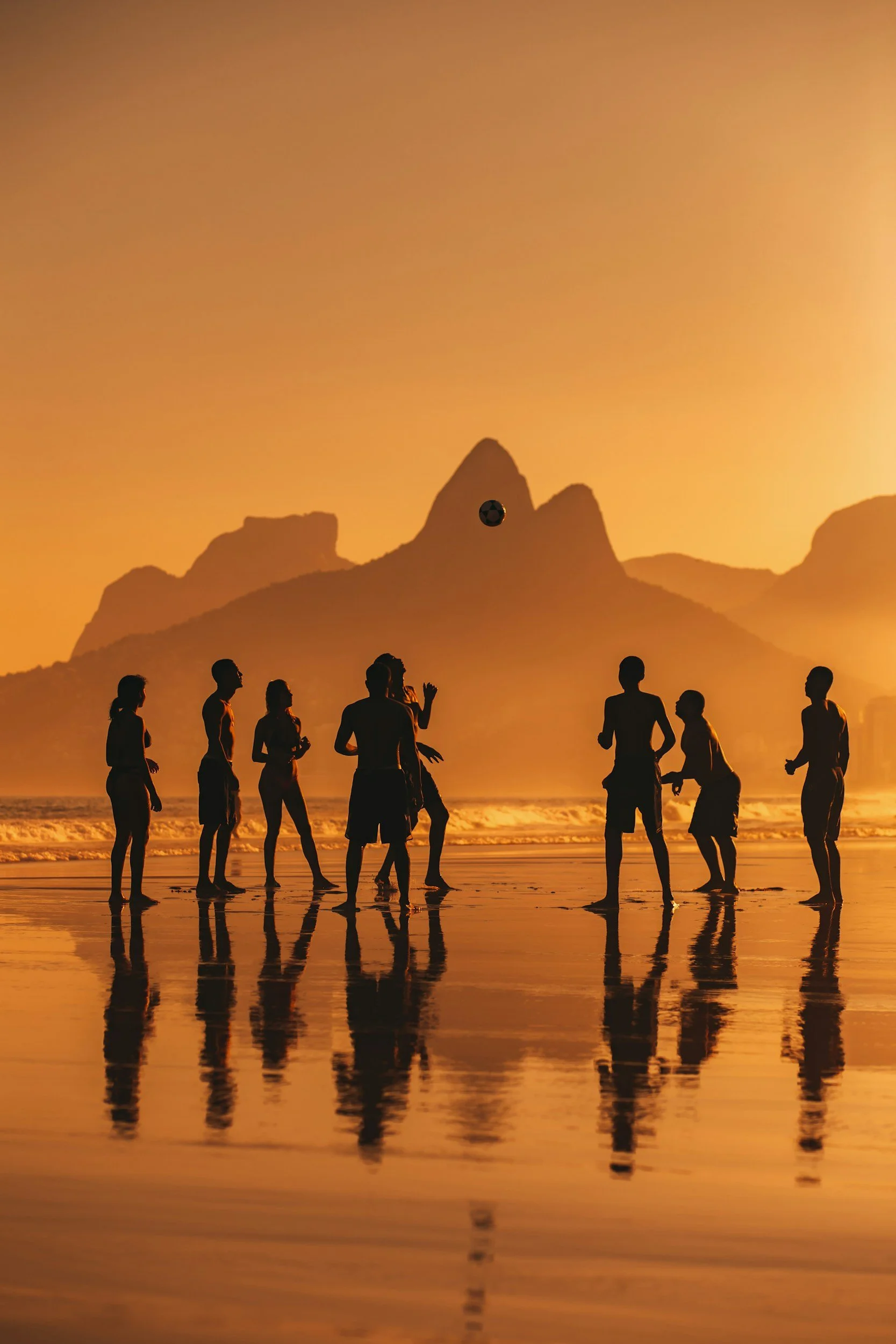 People on a beach during sunset, playing volleyball, with mountain silhouettes in the background and their reflections in the wet sand.
