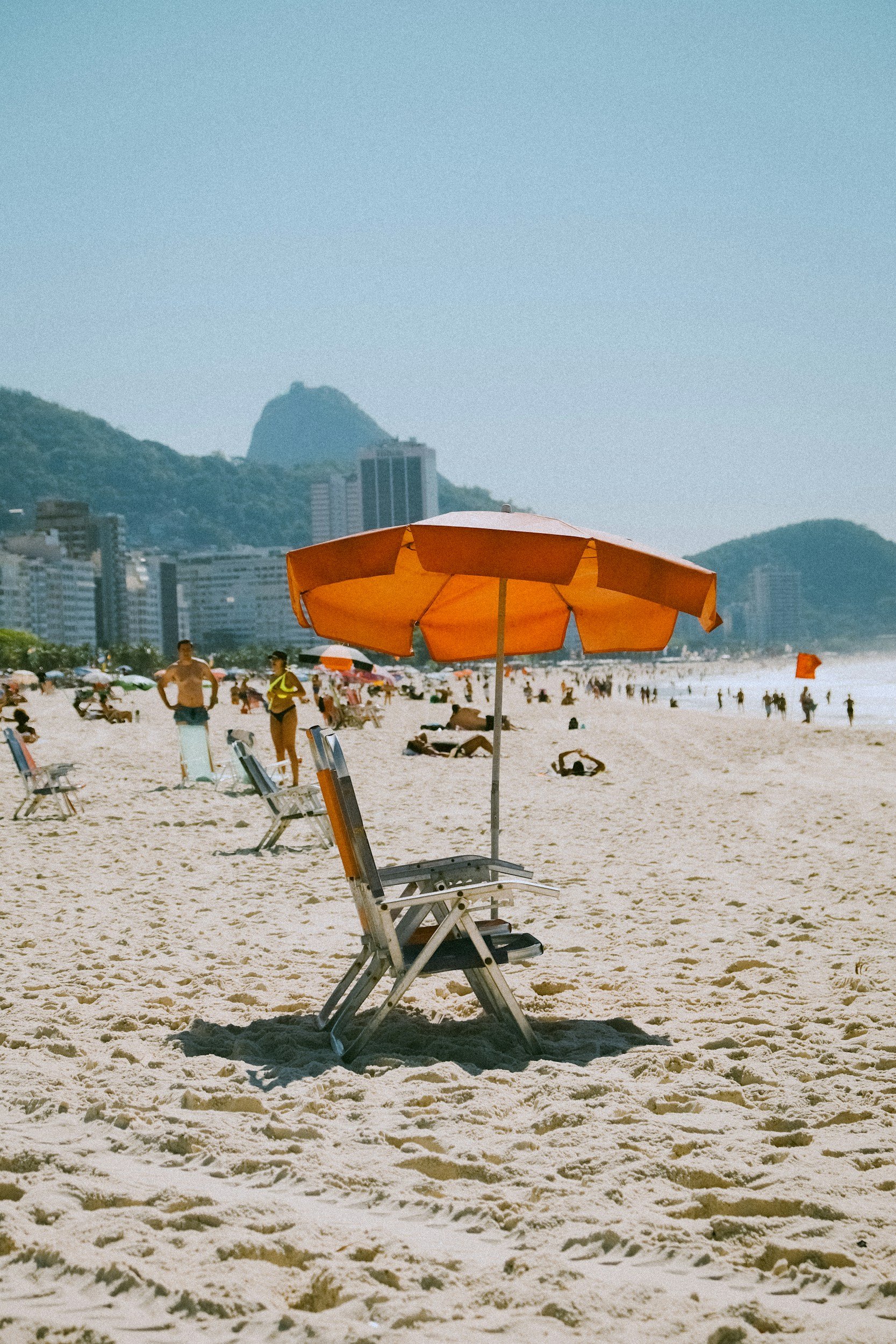 Empty orange beach umbrella with a gray lounge chair underneath on a sandy beach, with people in the background and tall buildings near the coast, with a mountain and a building on top in the distance.