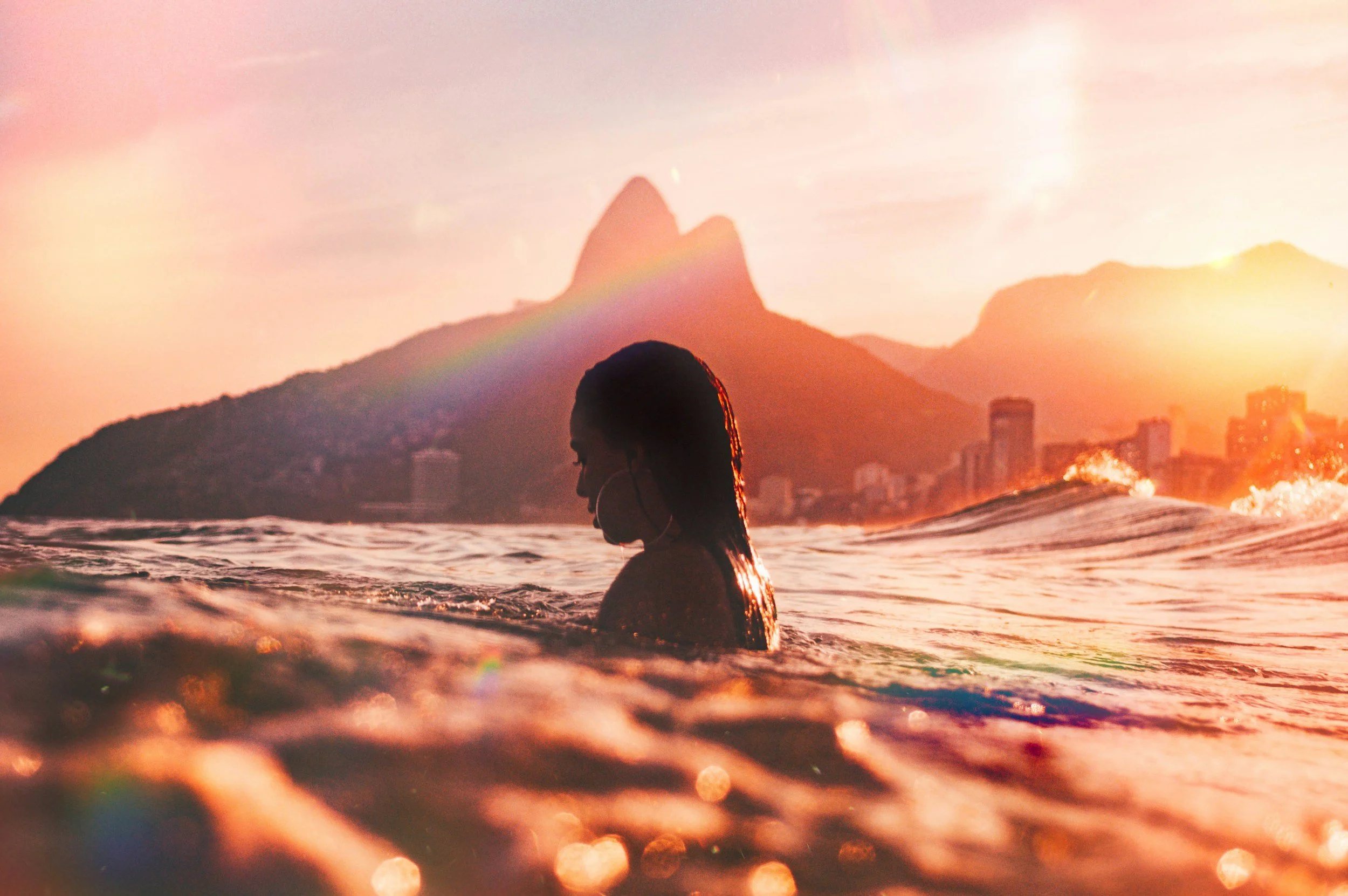 A person with long hair swimming in the ocean at sunset with a city skyline and mountains in the background.