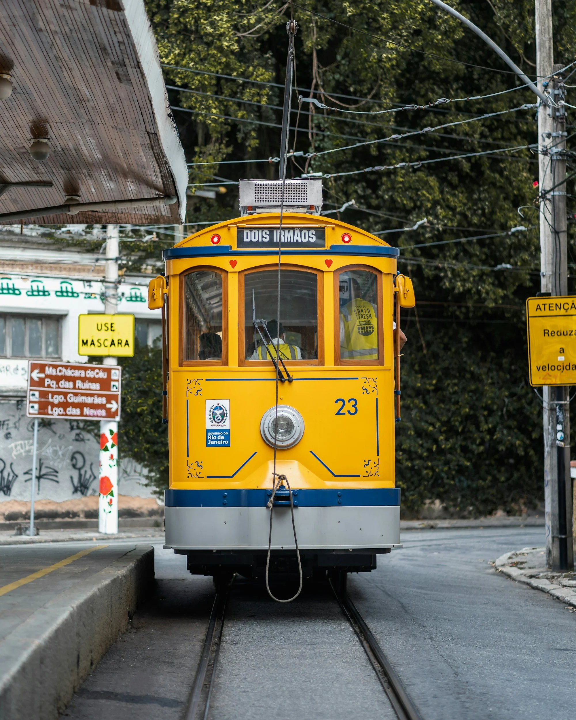 A yellow streetcar with a sign reading 'Dois Irmãos' on the front, traveling on tracks with overhead electric wires, passing under a metal awning, with trees and street signs in the background.