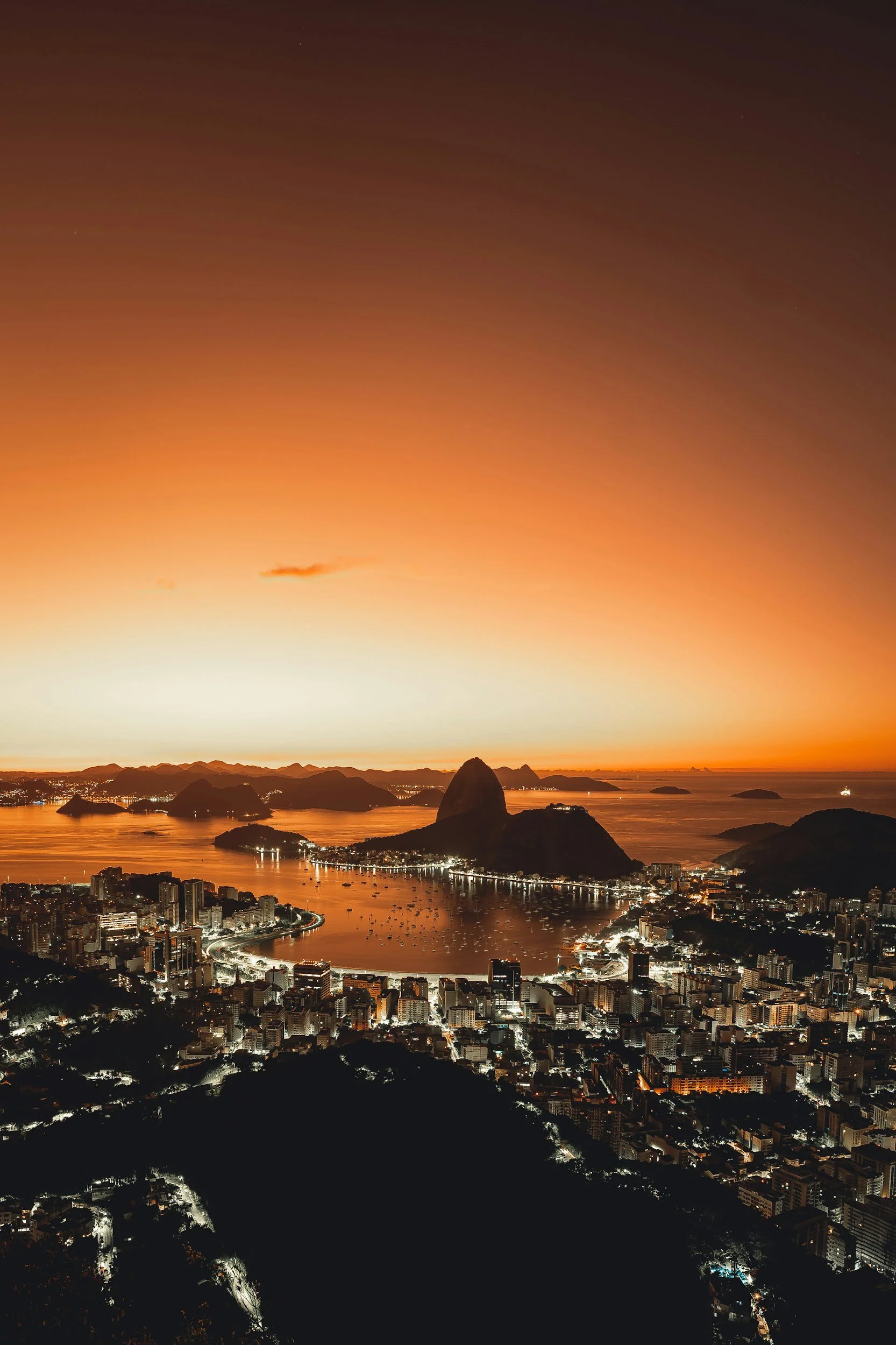 A cityscape at sunset with mountains, water, and buildings, including Sugarloaf Mountain in the background and a harbor with ships.