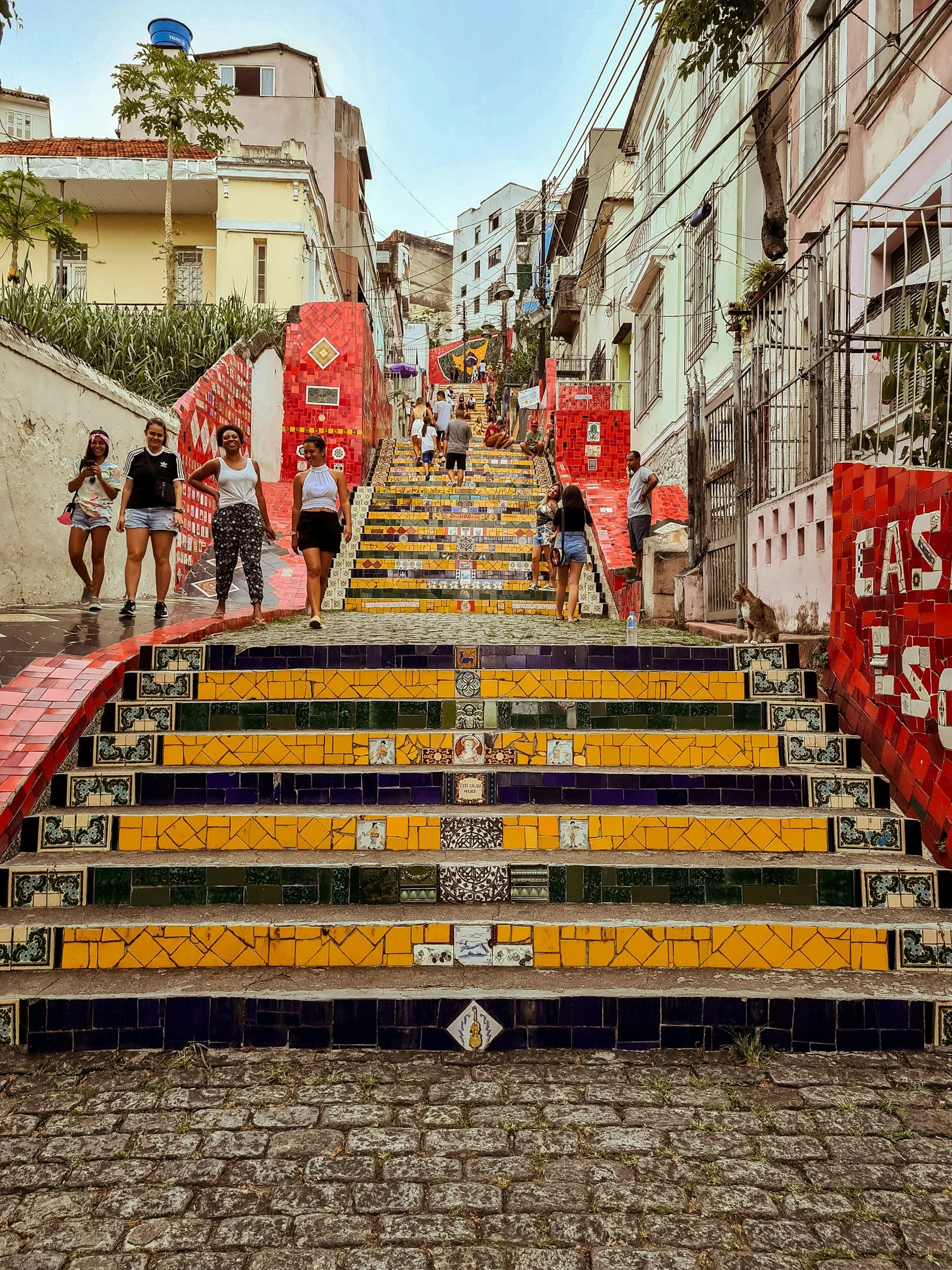 Colorful tiled staircase with people walking and sitting on the steps, surrounded by buildings and trees in a hilly urban area.