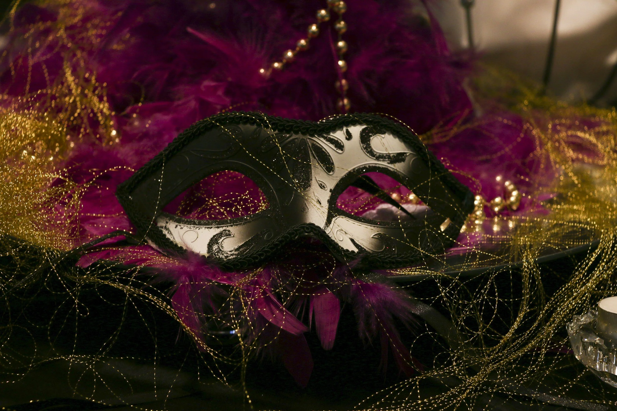 A decorative black and white masquerade mask adorned with feathers, surrounded by pink feathers, gold beads, and gold string decorations.