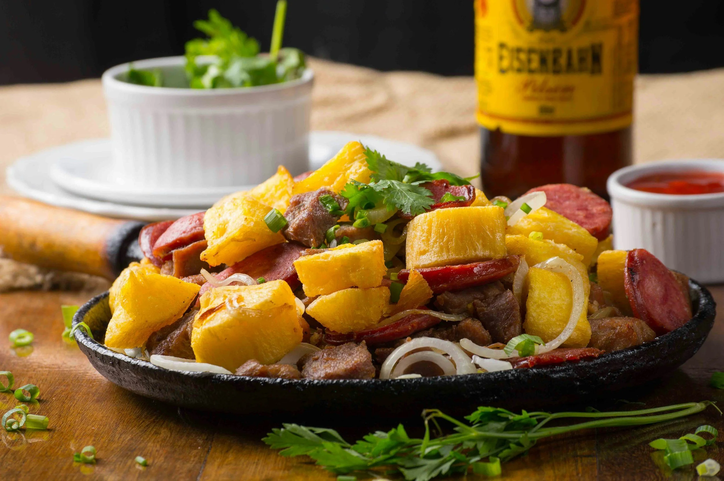 Cast iron skillet with a loaded breakfast hash featuring potatoes, sausage, and onions, garnished with fresh cilantro, on a wooden surface with scattered green onions and parsley, and a cup of salsa in the background.