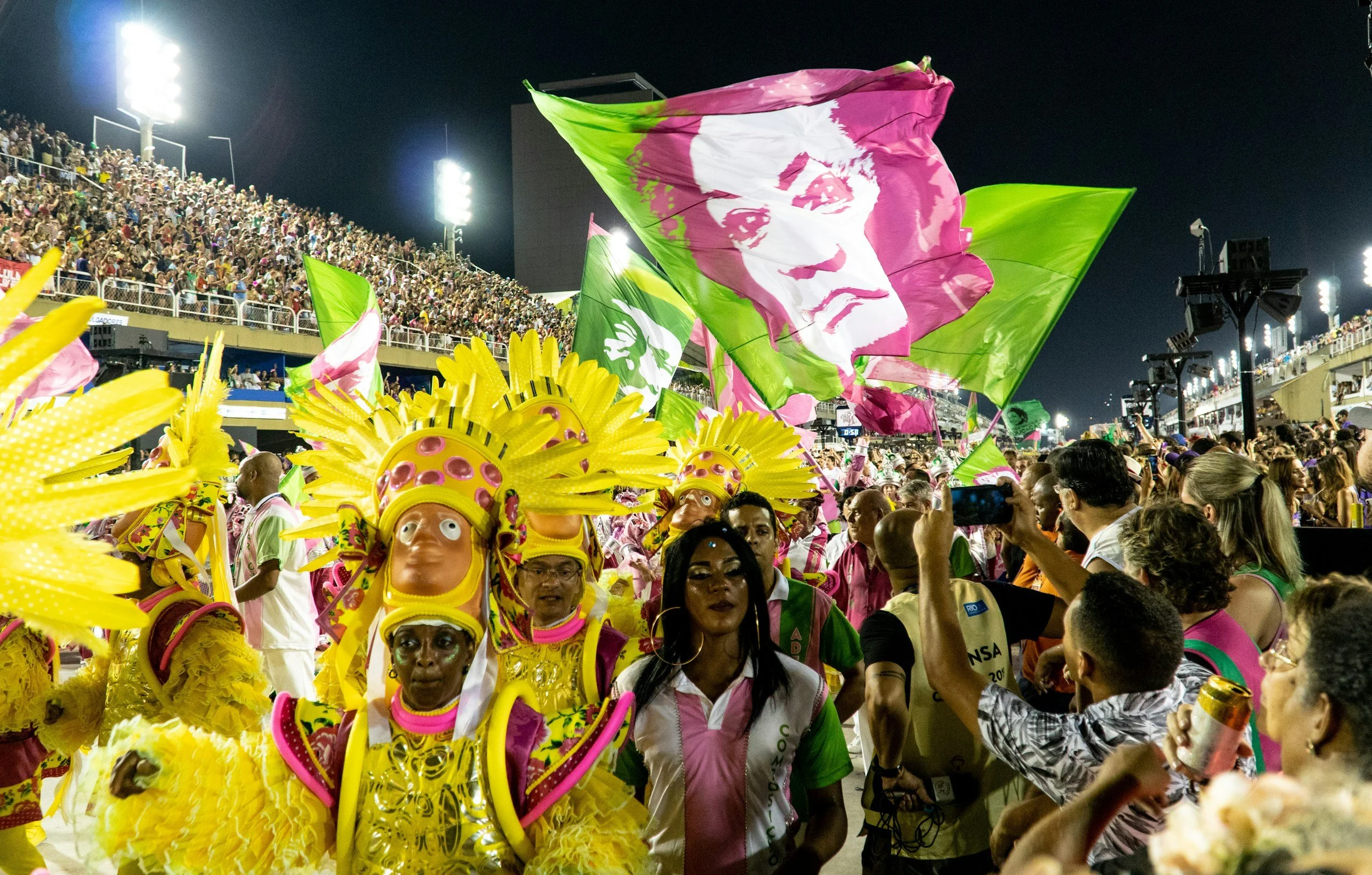 Carnival parade at night with performers in yellow costumes and giant flags, large audience on stadium sides, bright floodlights illuminating the scene.