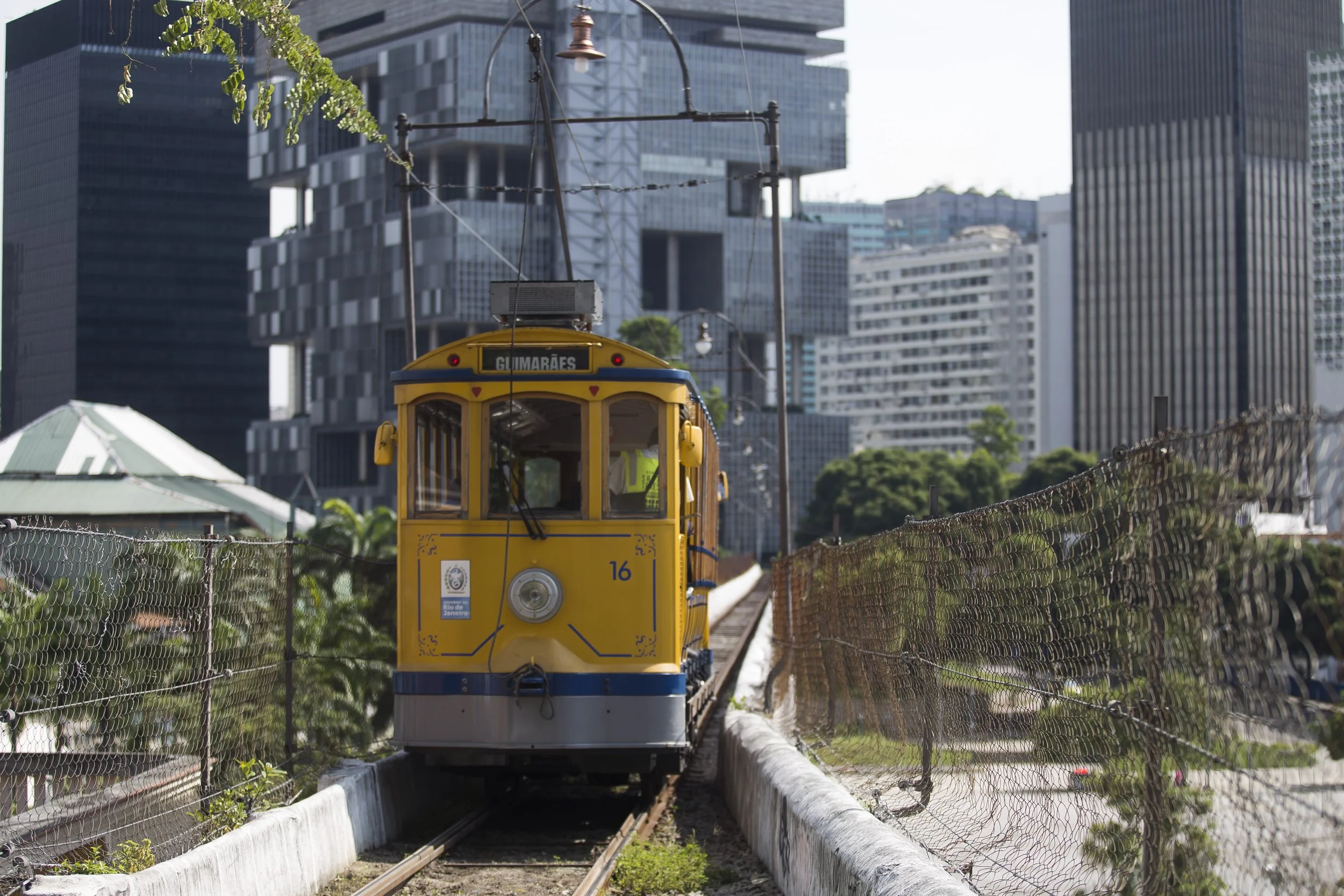 Yellow tram with the sign 'GILMARÈS' traveling on a narrow gauge railway track between a chain-link fence and concrete barriers, in an urban area with tall modern buildings in the background.