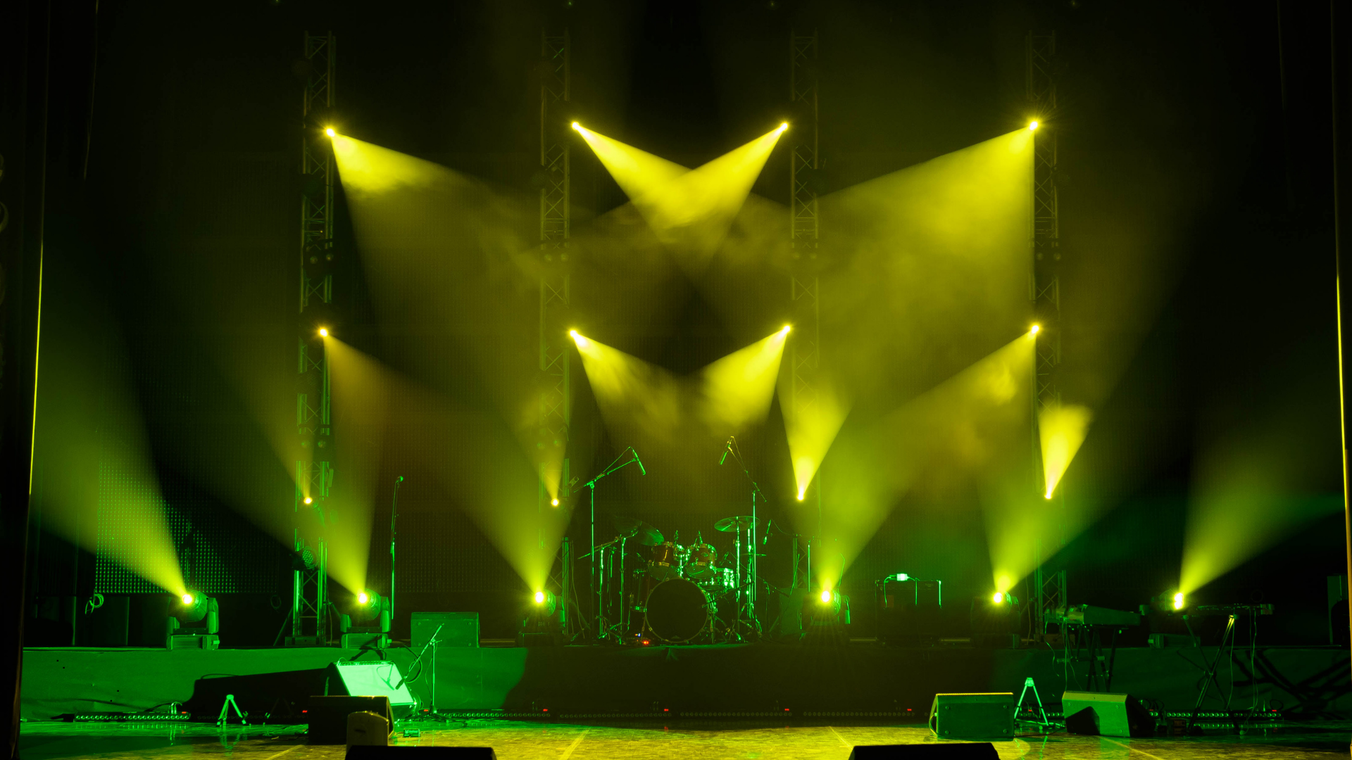 Stage with yellow and green stage lights, musical instruments including drums, microphones, and stands, set up for a concert or performance.
