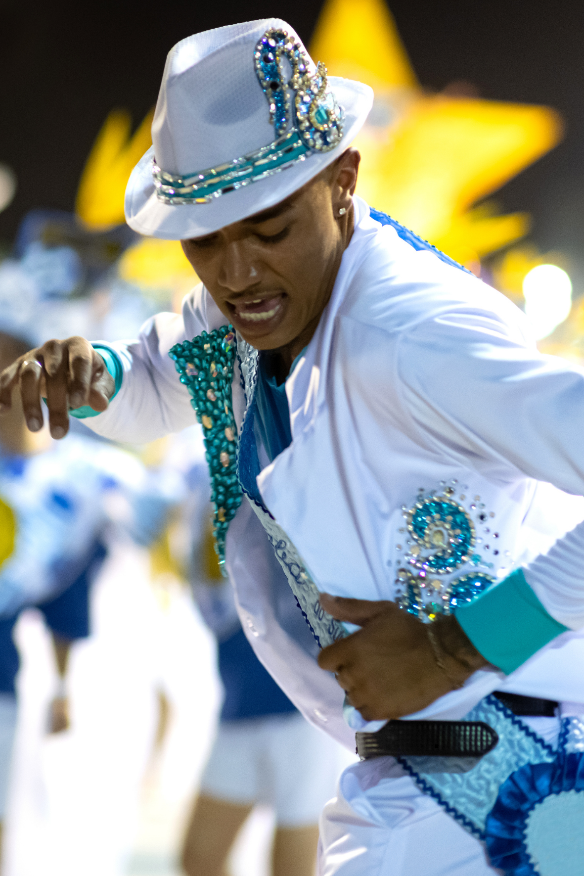 A man dressed in elaborate white and blue costume decorated with sequins and beads, wearing a white hat with blue embellishments, participating in a parade or performance at night with a large yellow star in the background.