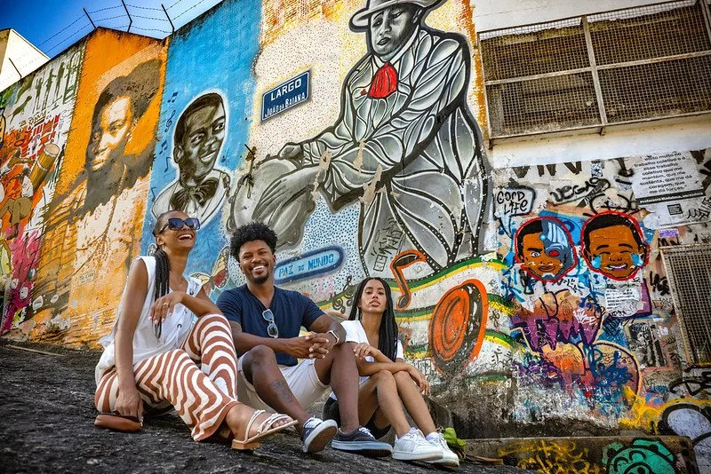 Three friends sitting in front of a colorful mural with portraits and graffiti art on a wall.