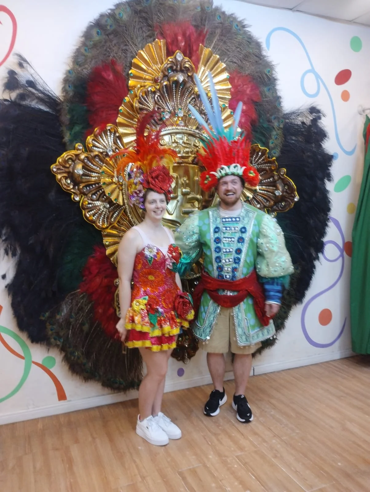 Two people in colorful costumes, standing in front of a decorative peacock feather and gold backdrop. The woman wears a vibrant red and orange dress with floral patterns, and the man wears a green and blue outfit with a feathered headdress.