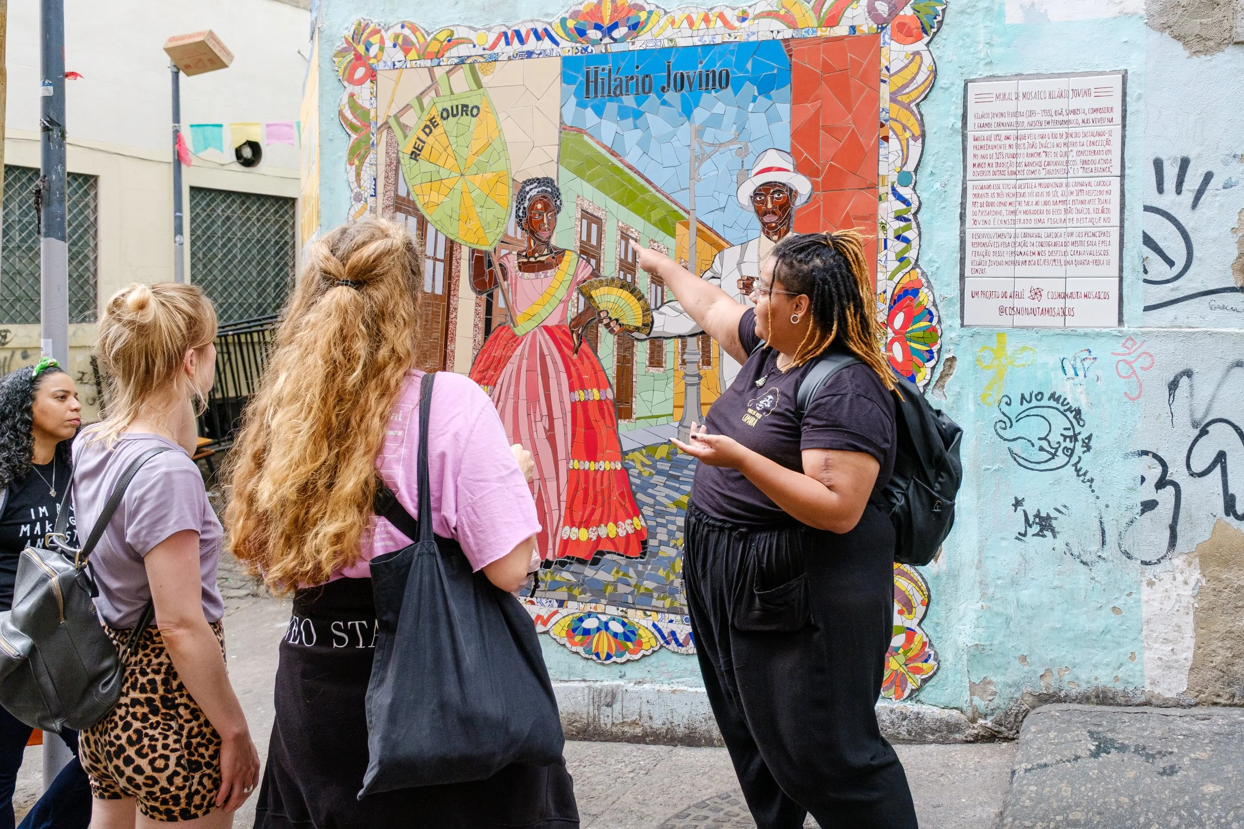A woman with dreadlocks and glasses is pointing at a colorful mosaic mural on a wall, explaining to a group of four young women who are attentively listening. The mural features two figures in traditional clothing, one with a fan and the other holdin