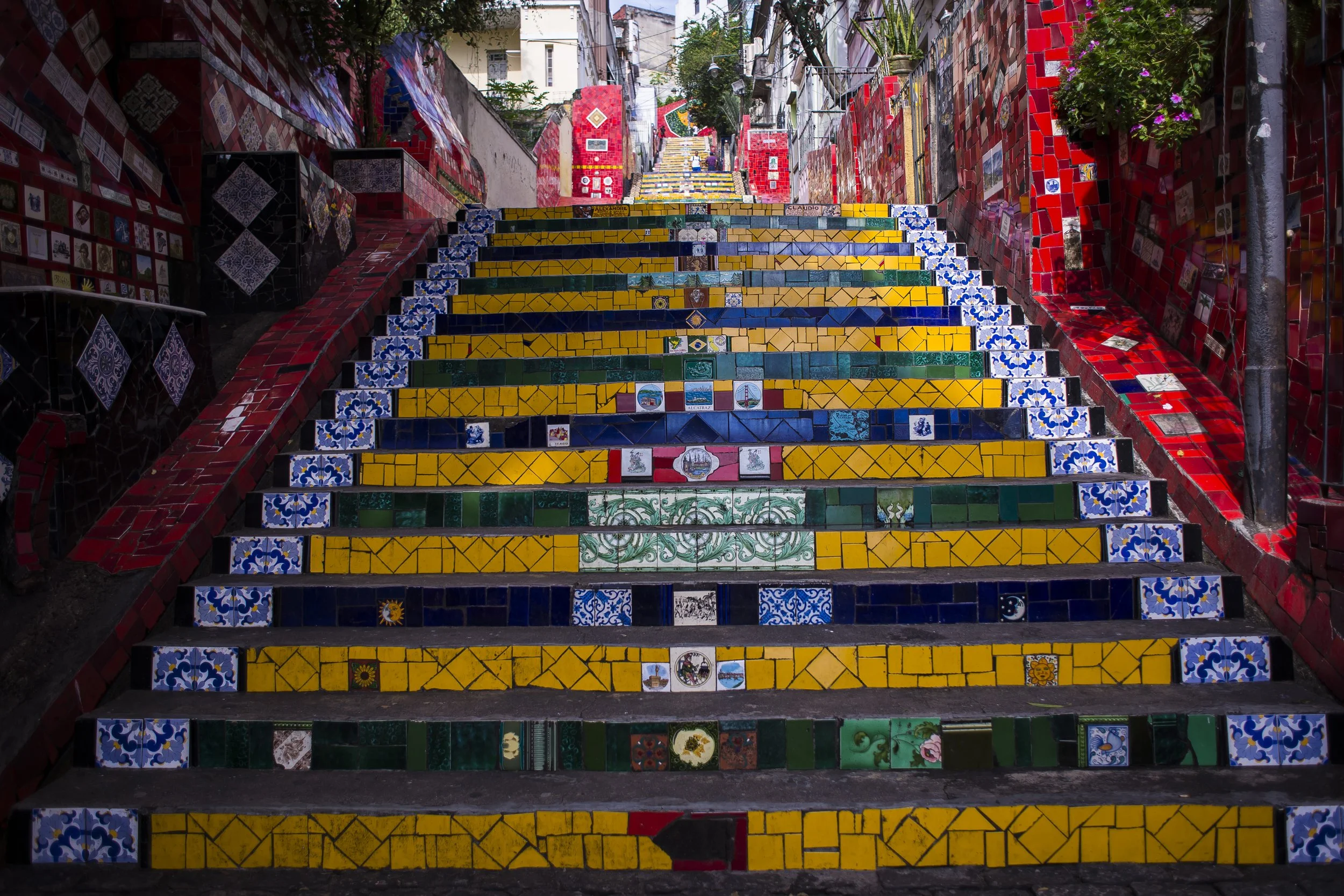 Colorful tiled stairs in a narrow outdoor alley, with walls decorated with various mosaic tiles and potted plants.