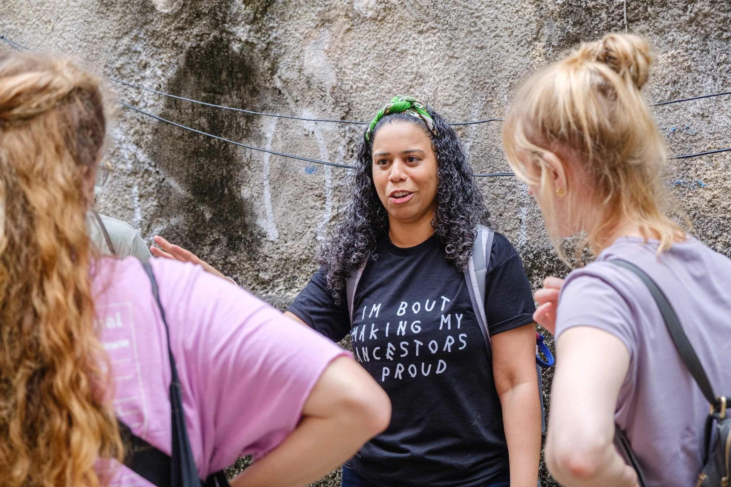 A woman with curly hair and a green headband talking to three women with red hair, among large rocks. The woman is wearing a black t-shirt with white text.