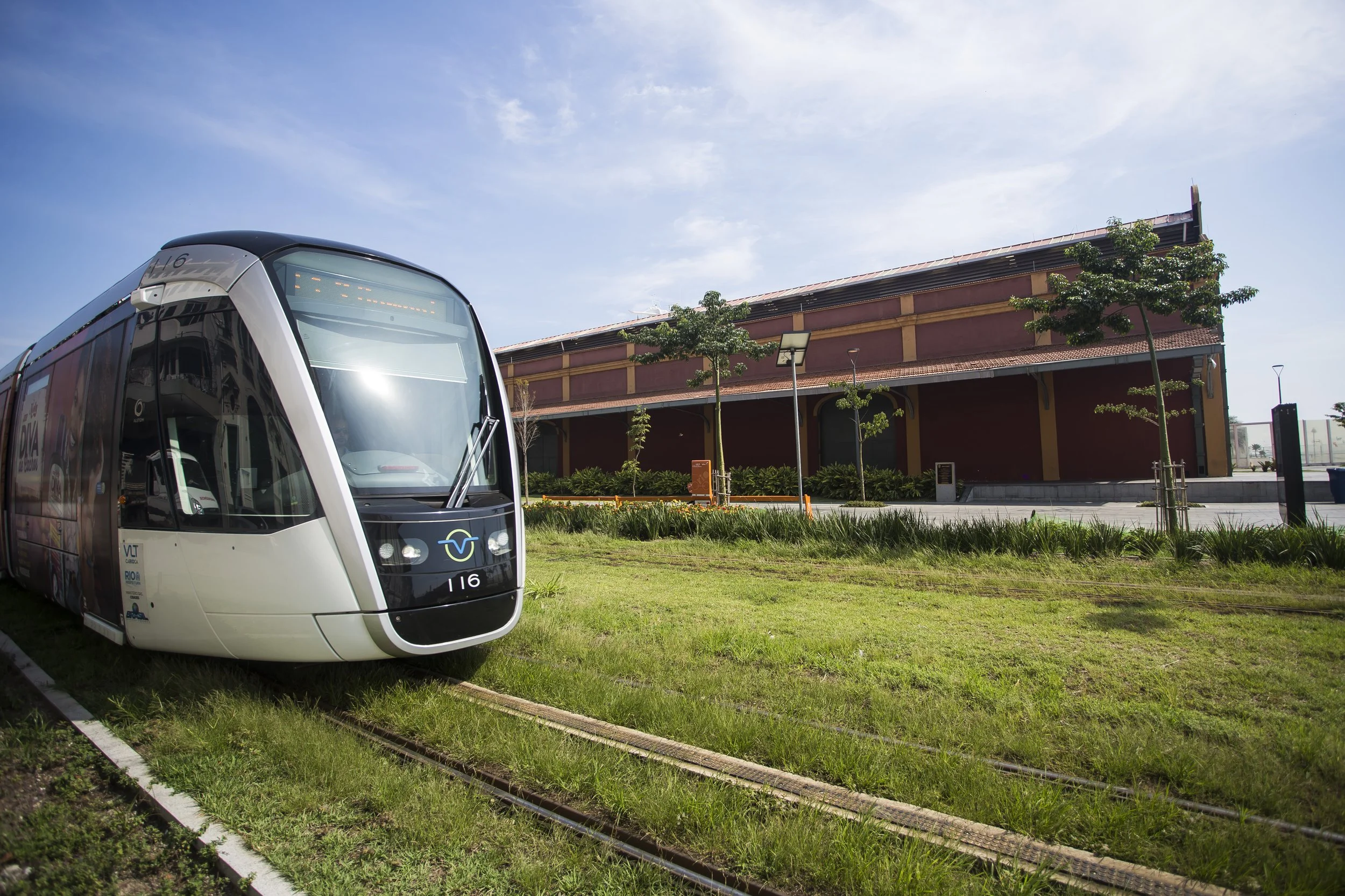 A modern white train with black accents on a grassy track beside a red and yellow building with trees and lampposts under a blue sky with some clouds.