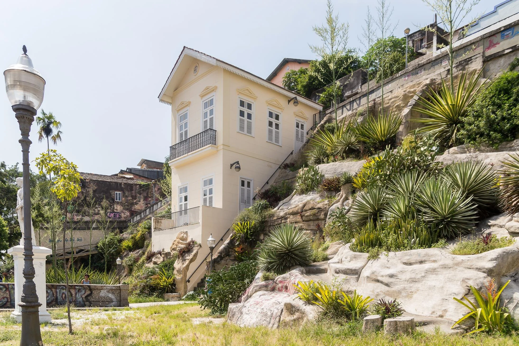 A multi-story yellow house with white windows and black wrought iron balcony, situated on a rocky hillside with lush green plants, palm trees, and a streetlamp in foreground.