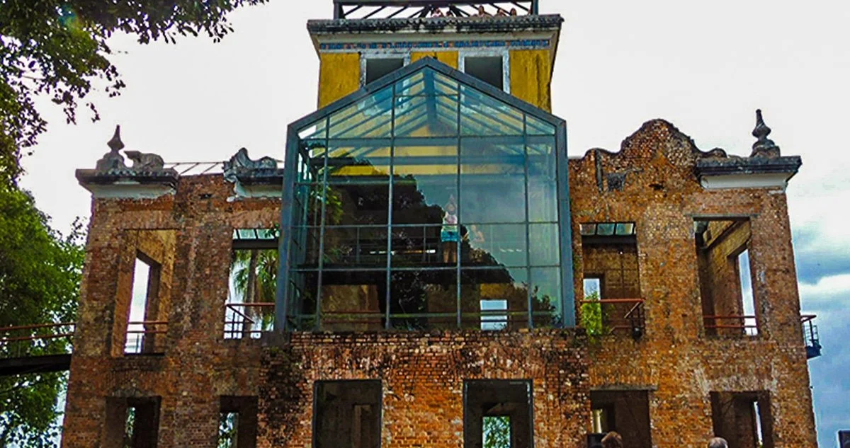 An old brick building with a modern glass extension on top, surrounded by green trees, under a cloudy sky.