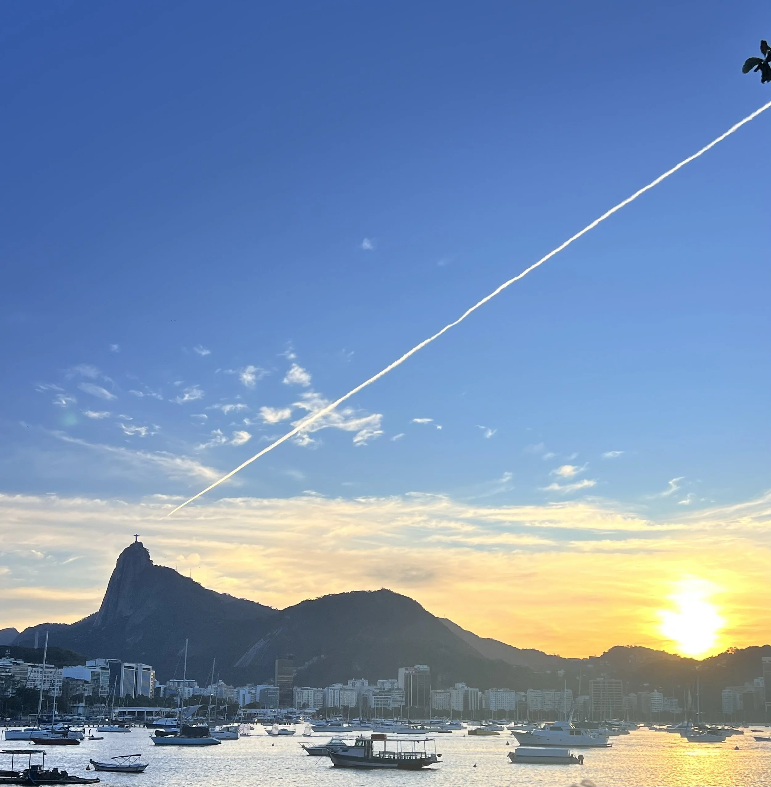 A marina filled with boats underwater, with a cityscape and mountains in the background, and a bright blue sky with some clouds and a contrail.