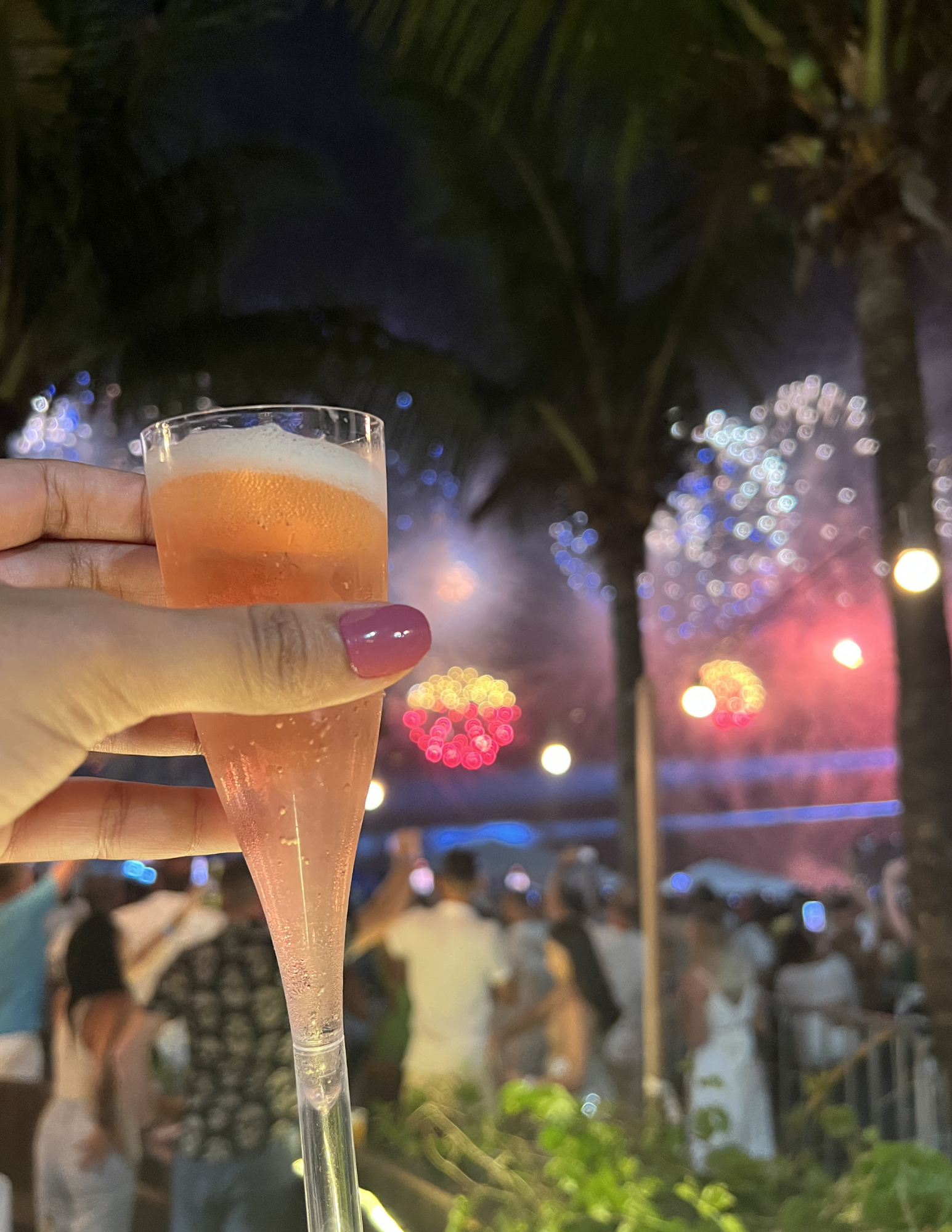 A person holding a pink cocktail in a glass with a straw at an outdoor event during nighttime, with blurred colorful lights and palm trees in the background.