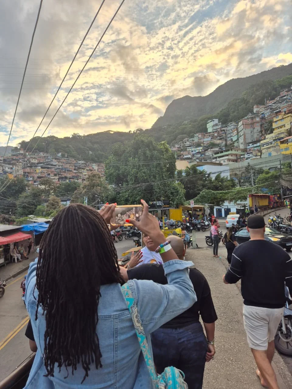 People walking along a street with a hillside covered with colorful houses in the background, under a sky with clouds and sunlight.