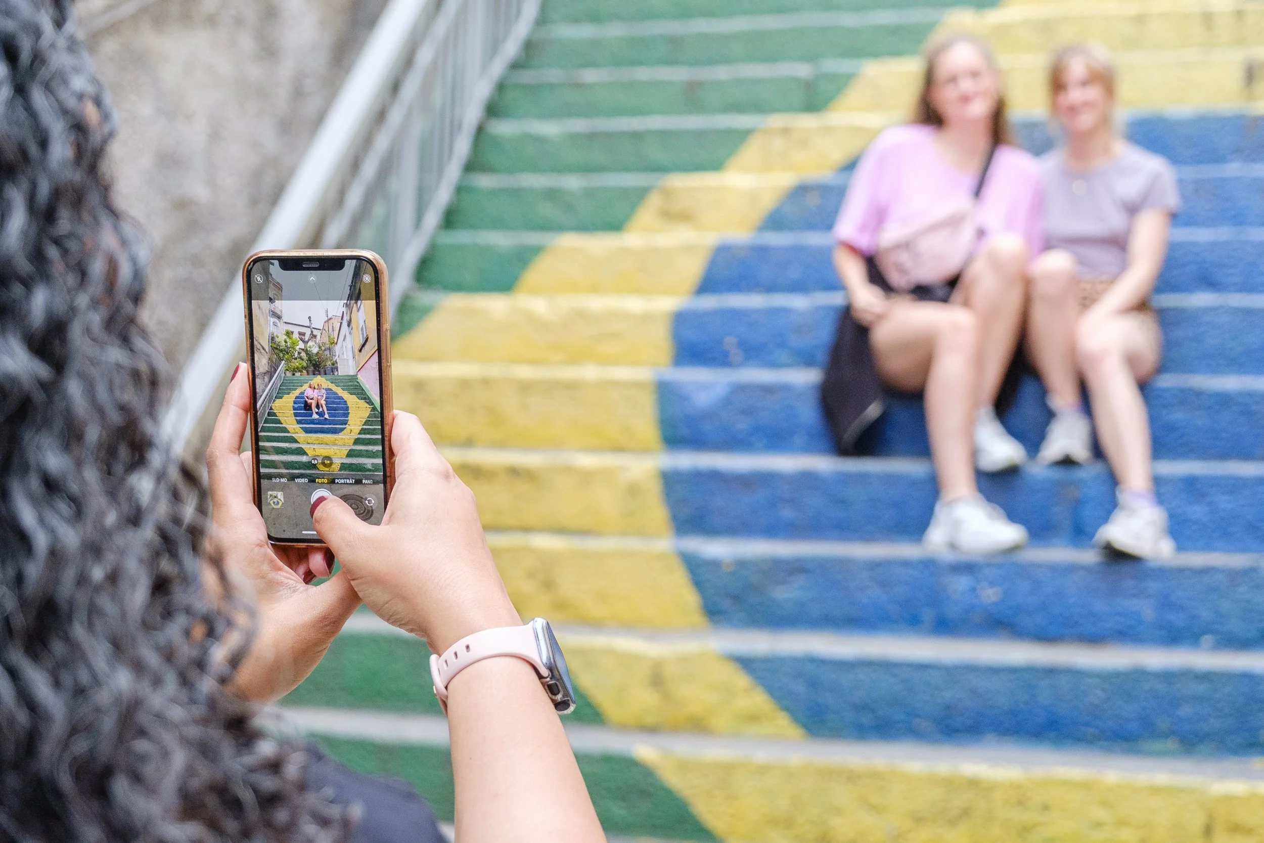 A person with curly hair taking a photo of two girls sitting on colorful outdoor stairs.