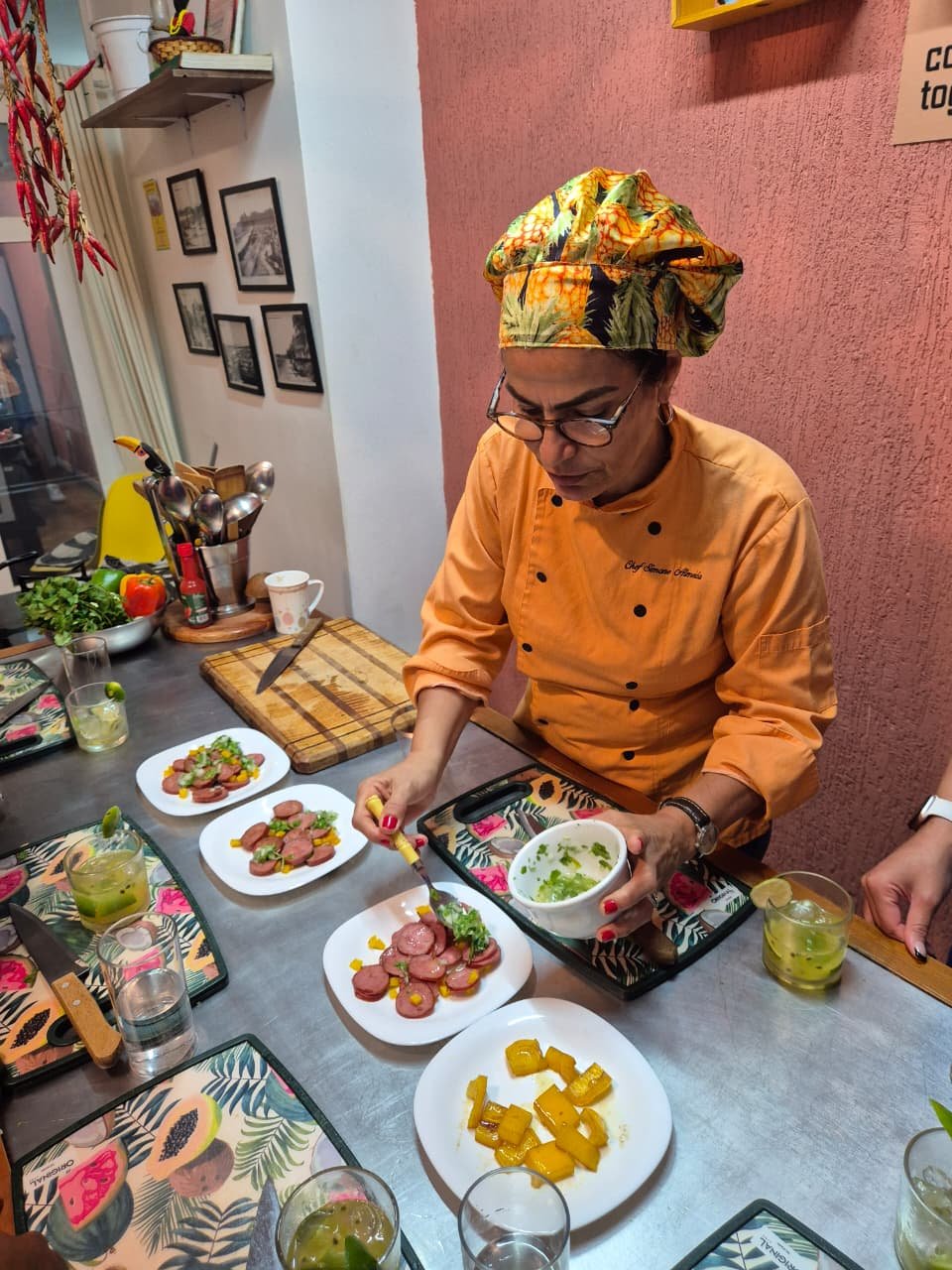 A chef prepares dishes in a kitchen, with plates of sliced meat topped with greens and garnishes on the table, along with drinks and utensils.