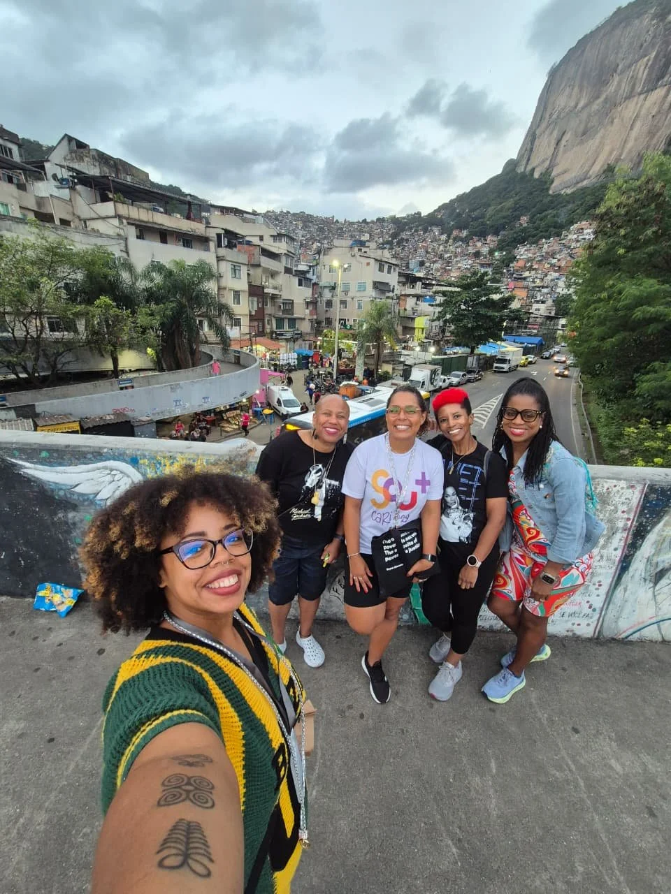 A group of five women standing on a bridge in Rio de Janeiro, Brazil, with the city's hillside and buildings in the background.