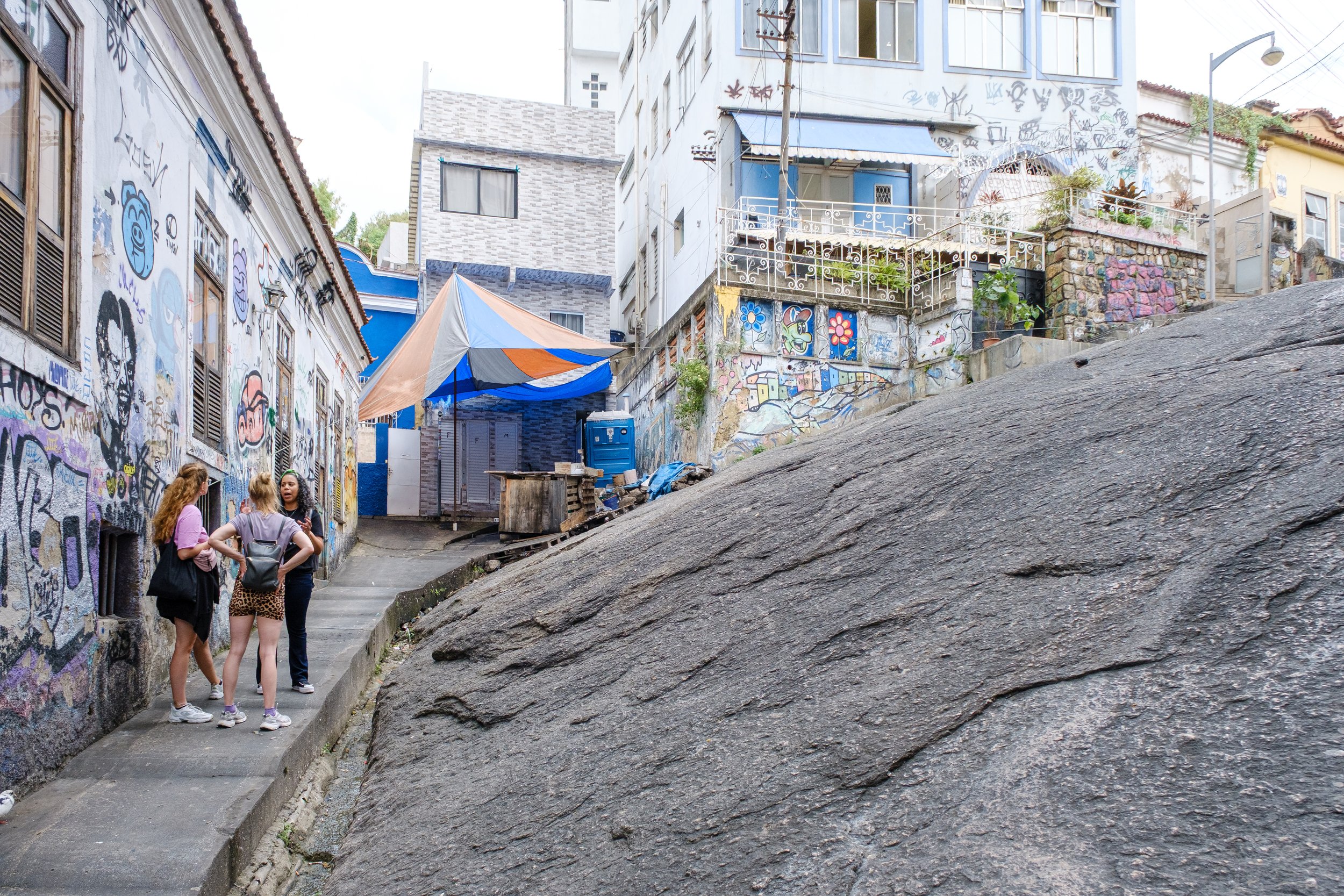 Three women standing on a narrow sidewalk in an urban area with buildings covered in graffiti; a large rock is in the foreground, and there are murals, a blue portable toilet, and a colorful umbrella in the background.