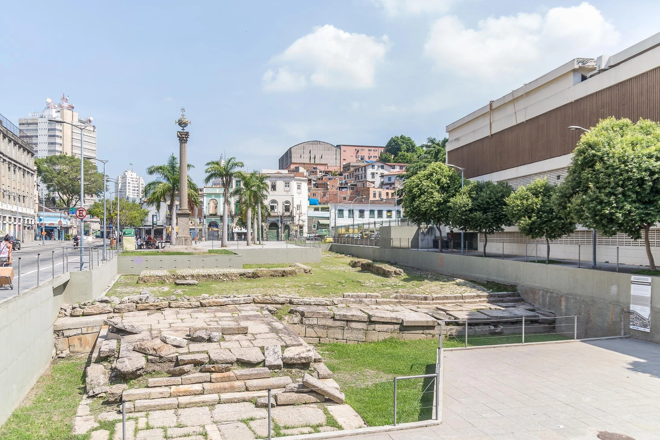 Ancient stone steps and foundations at an archaeological site in a city park, with modern buildings, trees, and a blue sky with clouds in the background.