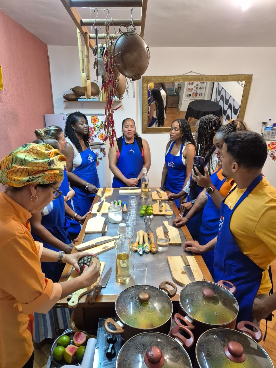 Group of people in a cooking class gathering around a wooden table with cooking utensils, ingredients, and a pineapple, in a kitchen with hanging pots and a mirror.