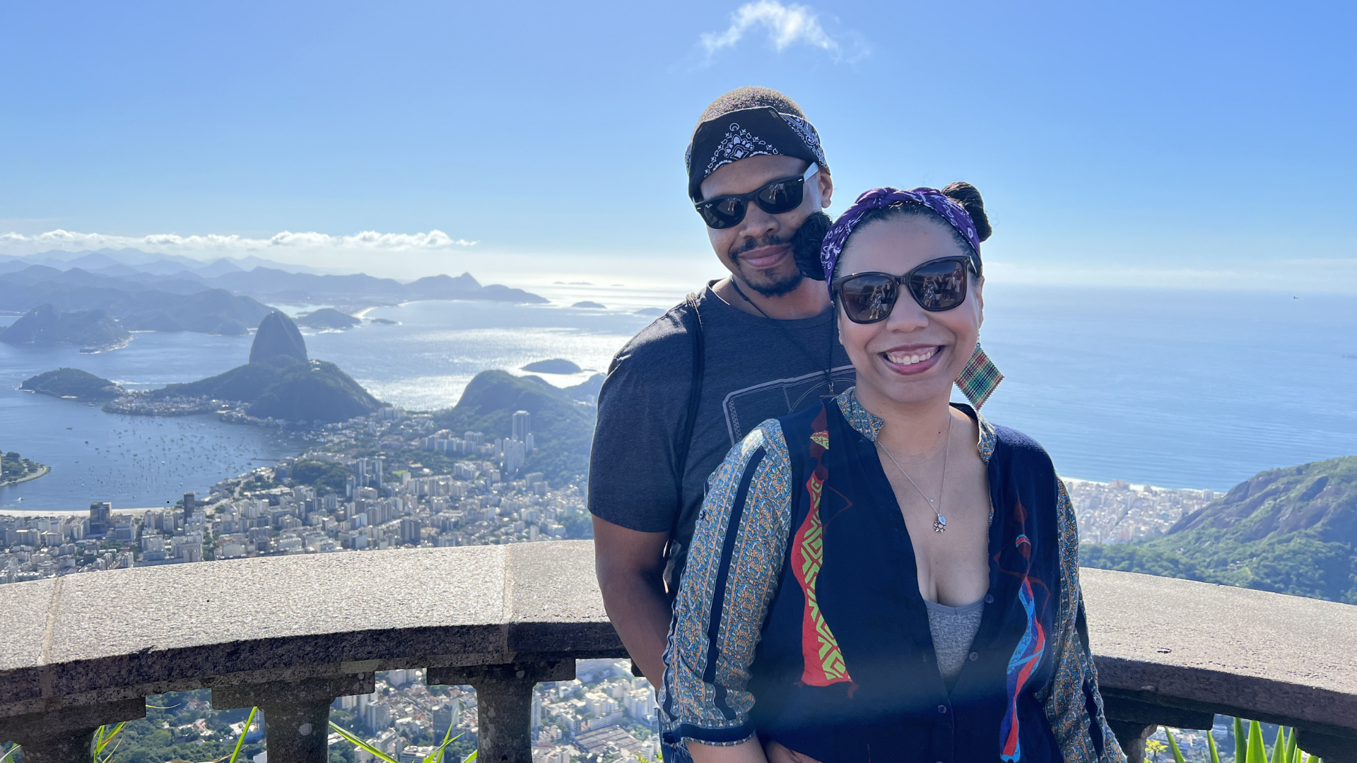 Two smiling women wearing sunglasses and headscarves stand on a viewing platform overlooking Rio de Janeiro with Sugarloaf Mountain and the city below during daytime.