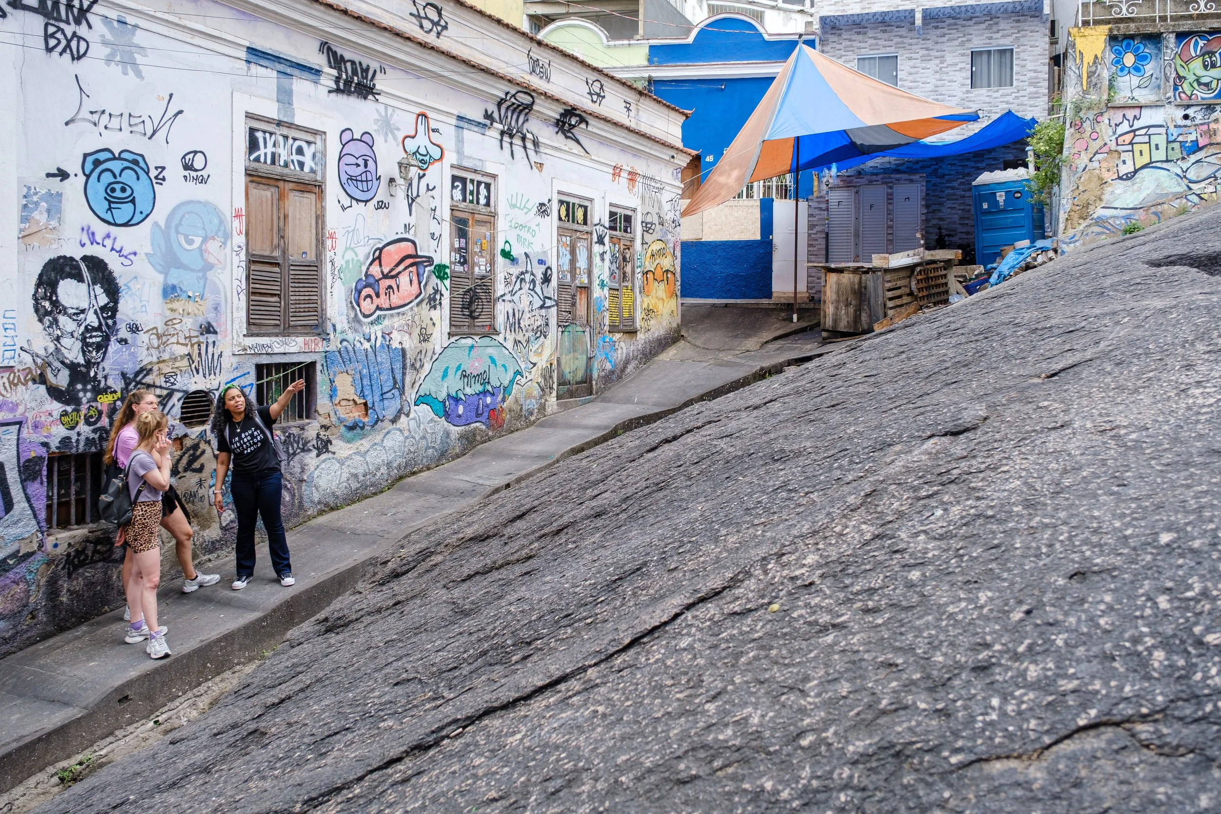 A group of three women standing on a sidewalk next to a graffiti-covered wall, with a rolling hill in the foreground. The graffiti features cartoon characters and various colorful tags. There is a building with windows behind the women and an umbrell