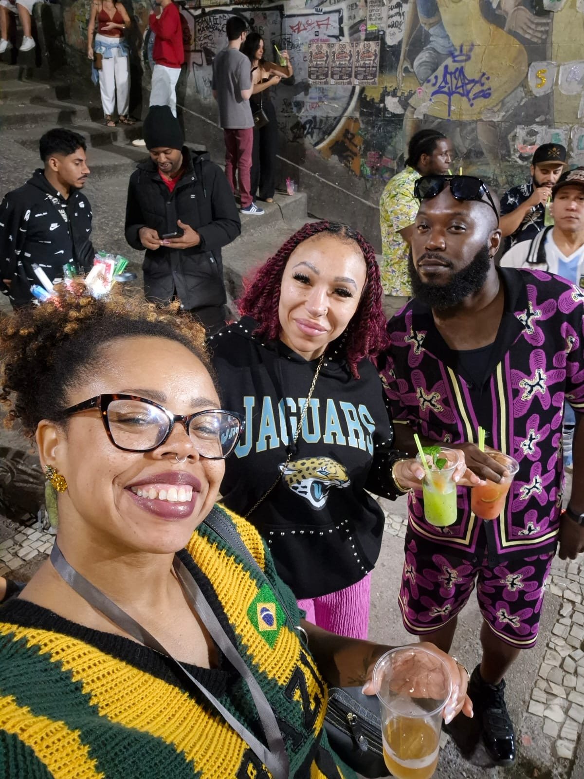 A group of diverse friends taking a selfie at a lively outdoor event, holding drinks, smiling, and dressed in colorful casual attire. In the background, other people are socializing and graffiti art decorates the wall.