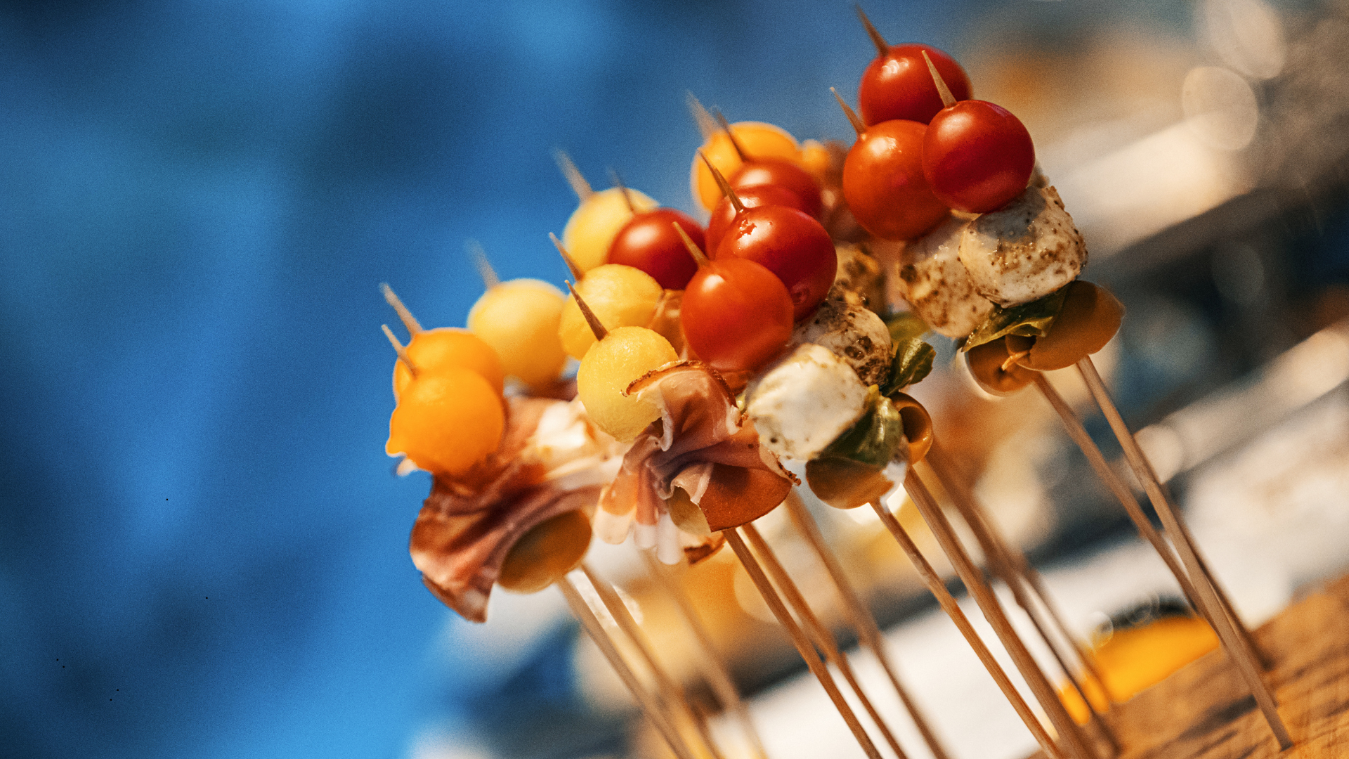 Close-up of skewers with small grilled tomatoes, mozzarella cheese, and herbs on a grill rack, with a blurry blue background.