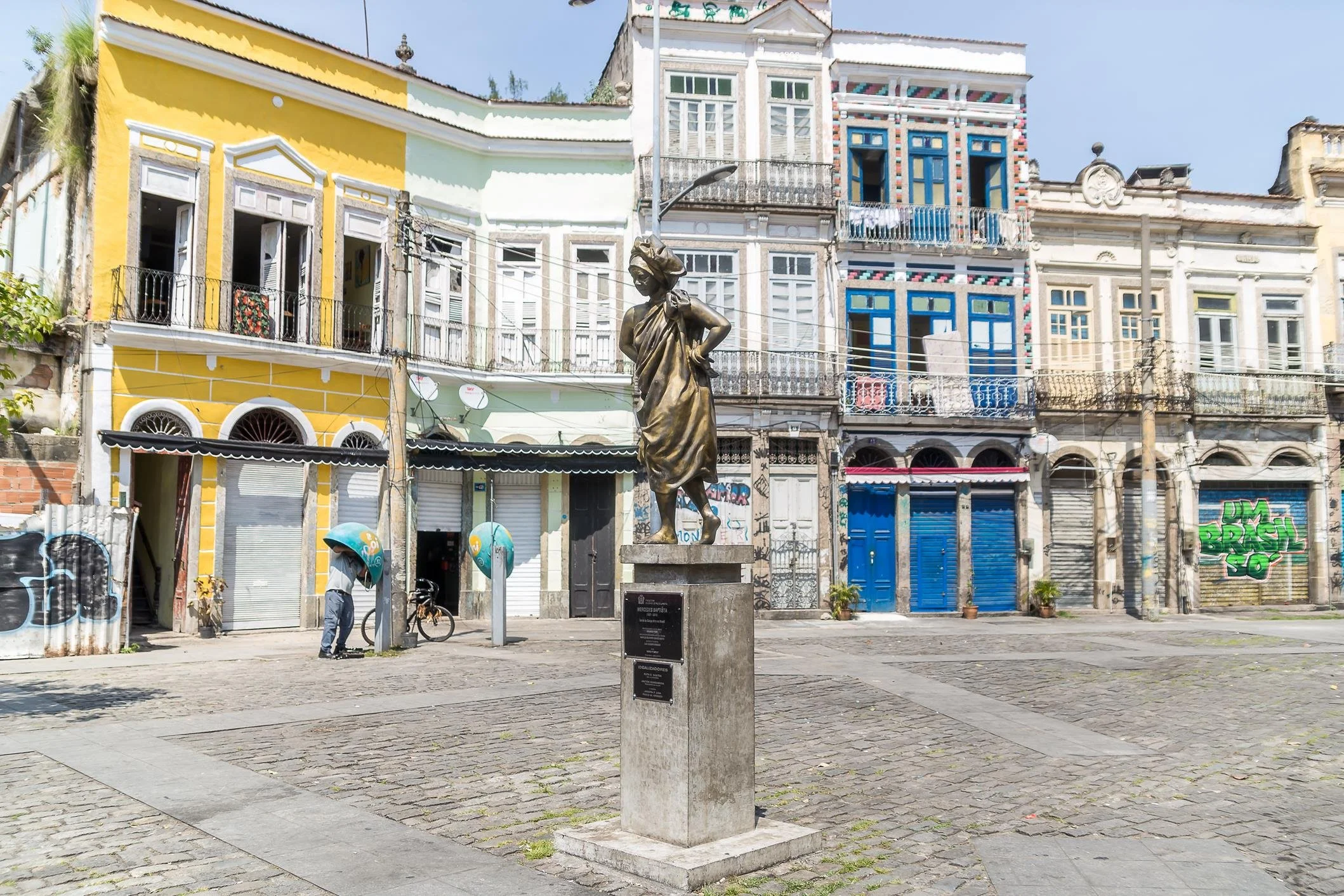 A city square with colorful colonial-style buildings, a bronze statue of a woman in traditional clothing, and a stone plaque on a pedestal. People are walking and cycling, and graffiti is visible on some walls.