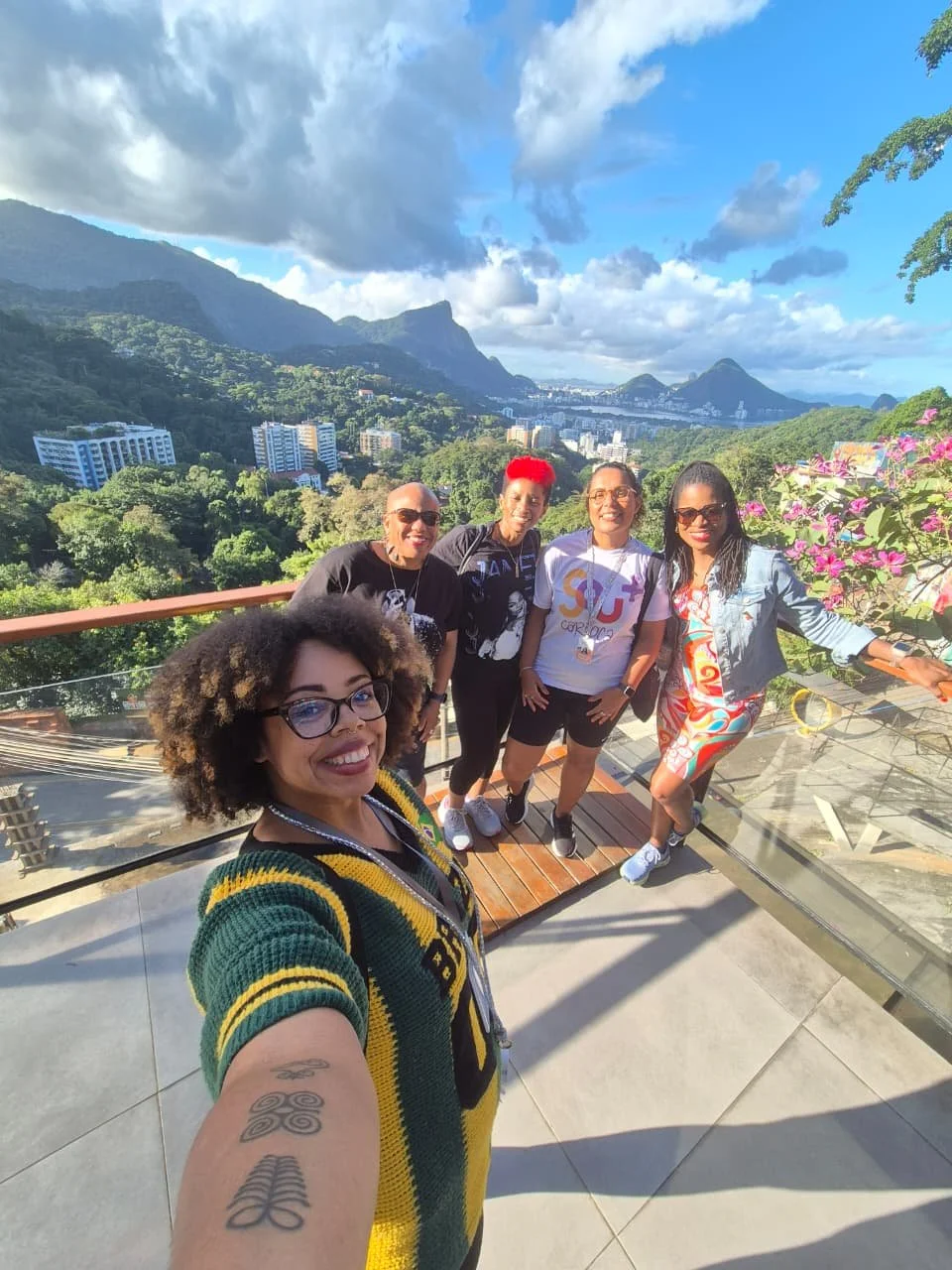 Group of five women taking a selfie on a balcony with mountains and lush greenery in the background, bright blue sky, and some clouds.