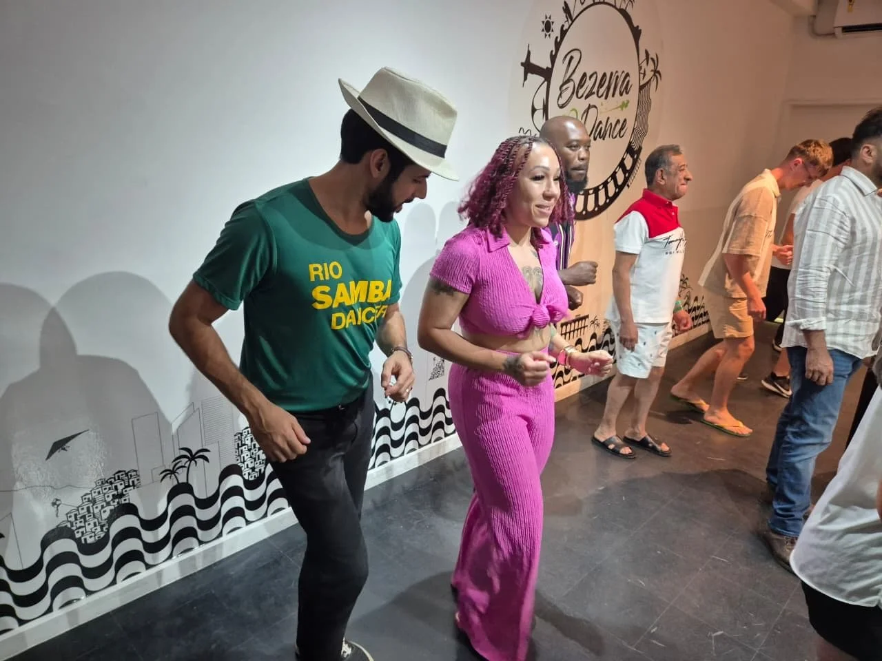 People dancing in a line at a dance studio with a sign in the background that reads 'Bezerra Dance'.